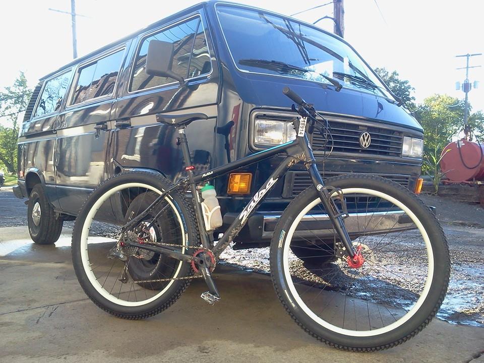 Salsa Mukluk: A black Volkswagen van parked on a driveway with a mountain bike positioned in front. The bike has distinctive white tires and a water bottle attached to its frame. Sunlight illuminates the scene, highlighting the vehicle and bike against a backdrop of greenery and utility poles.