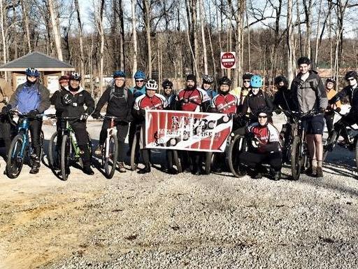 A group of cyclists standing together with mountain bikes, holding a sign that reads “EMBRC.” The setting appears to be a gravel path in a wooded area, with trees in the background and a building visible to the side. The participants are wearing cycling gear and helmets, showing a mix of enthusiasm and camaraderie.