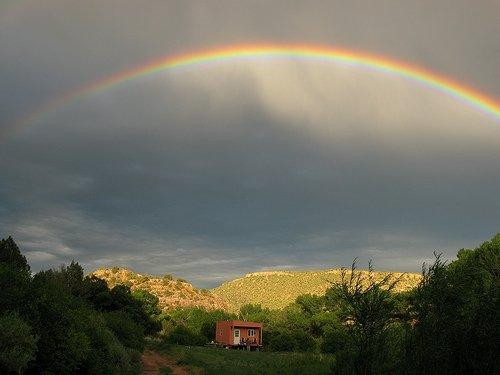 A vibrant rainbow arcs over a picturesque landscape featuring rolling hills and greenery, with a small house nestled in the foreground. The sky is partly cloudy, creating a dramatic backdrop for the colorful rainbow.