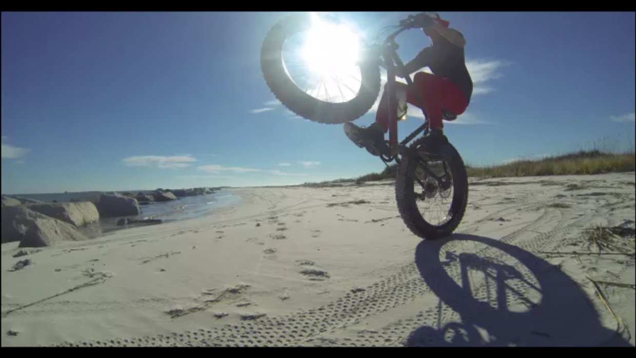 A person performing a wheelie on a fat bike on a sandy beach, with the sun behind them creating a halo effect. The scene features a clear blue sky, rocks along the shoreline, and tire tracks in the sand.