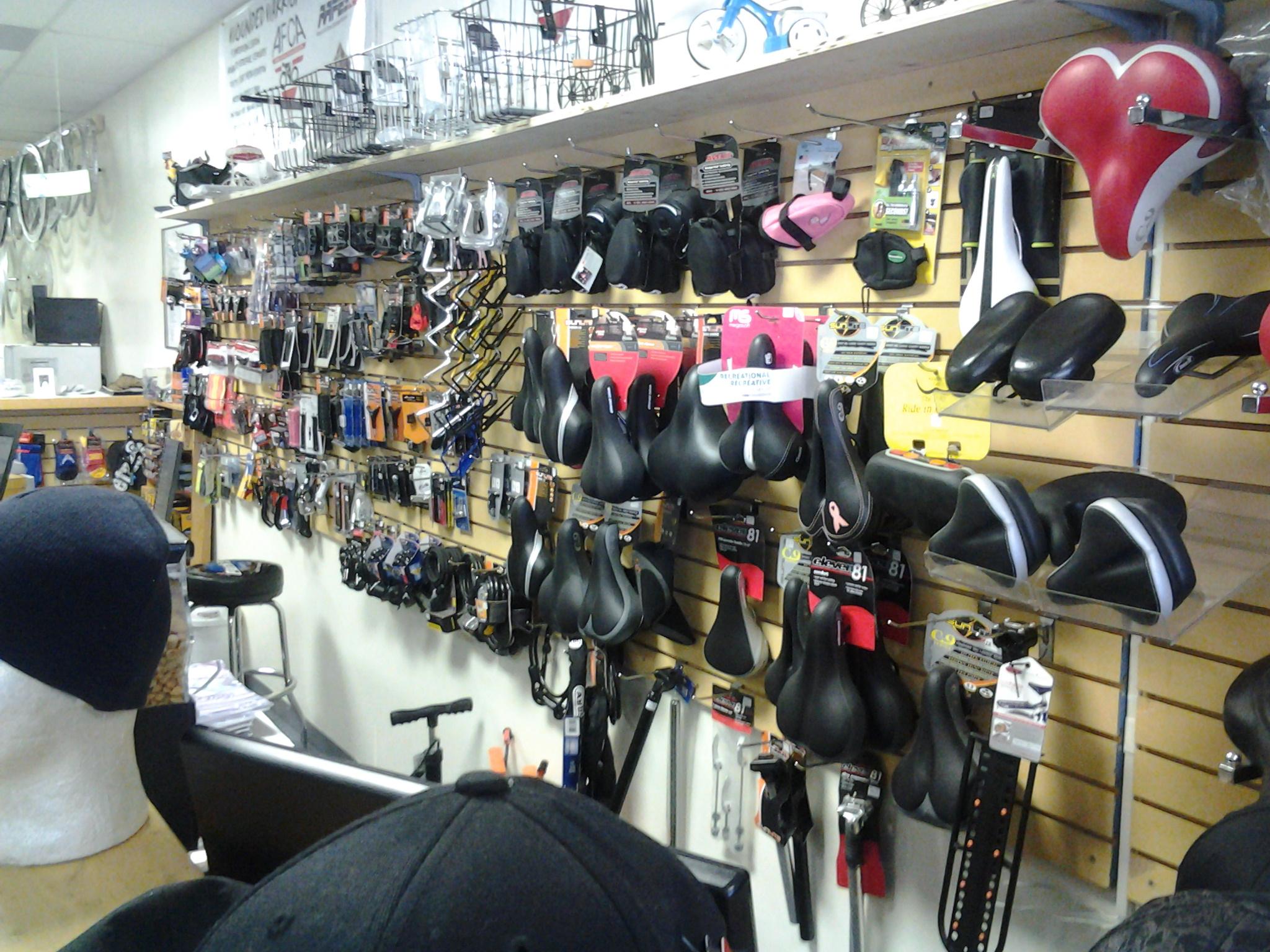 A display wall in a bicycle shop showcasing various bike seats and accessories. The wall features an array of different styles and colors of bicycle saddles, along with tools and other cycling gear arranged neatly. The shop has a friendly atmosphere, with a visible seat selection and additional merchandise in the background.