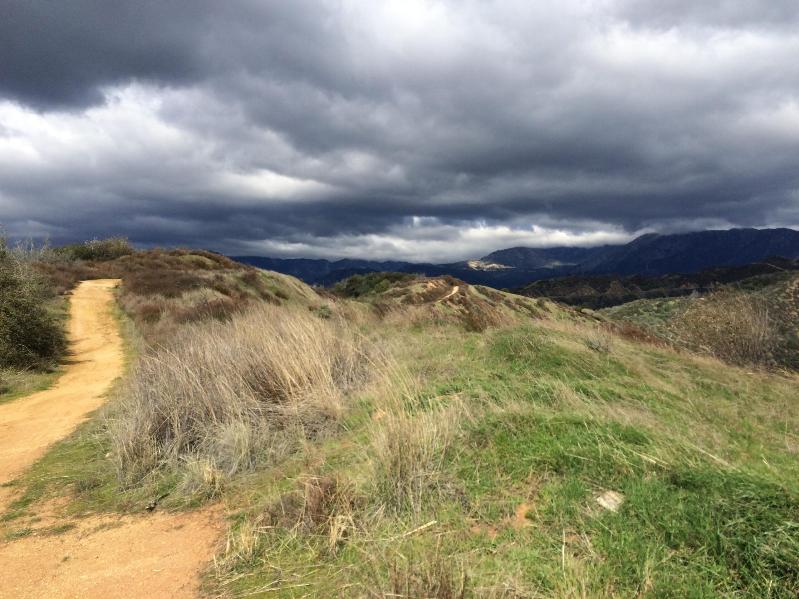 A winding dirt path leads through rolling hills covered in dry grass and shrubs, under a dramatic sky filled with dark, ominous clouds. In the background, mountains can be seen, creating a scenic yet moody landscape. Monroe Truck Trail mountain bike trail.