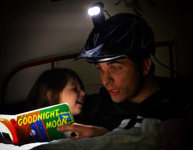A person wearing a helmet with a light is reading "Goodnight Moon" to a young child in a cozy, dimly lit room. The child is smiling and engaged, sitting close to the reader.