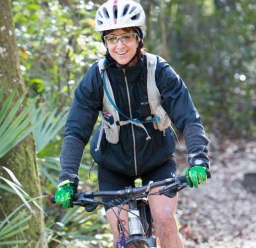 A smiling person riding a mountain bike on a wooded trail, wearing a helmet, sunglasses, and a black jacket with a hydration pack. They have green gloves and are surrounded by greenery.