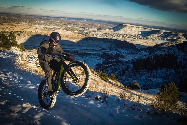 A person riding a fat tire bicycle performs a wheelie on a snowy trail, with a panoramic view of a valley and mountains in the background under a clear blue sky.