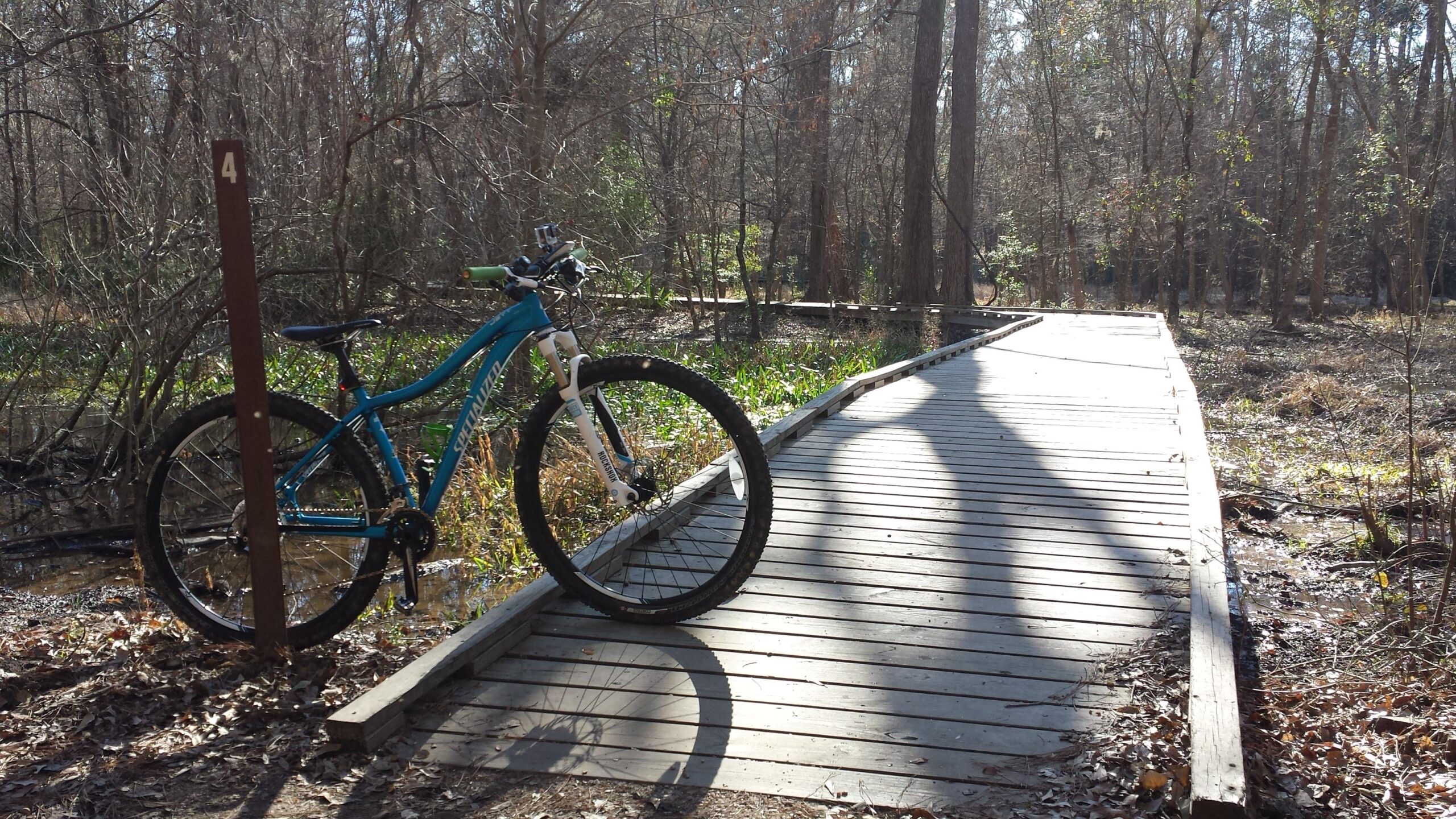 A blue mountain bike is parked beside a wooden boardwalk in a wooded area. The bike is positioned next to a post marked with the number 4. The scene features trees with bare branches and some green foliage at the edge of a water feature, with fallen leaves scattered on the ground. Sunlight filters through the trees, casting shadows on the boardwalk. Huntsville State Park mountain bike trail.