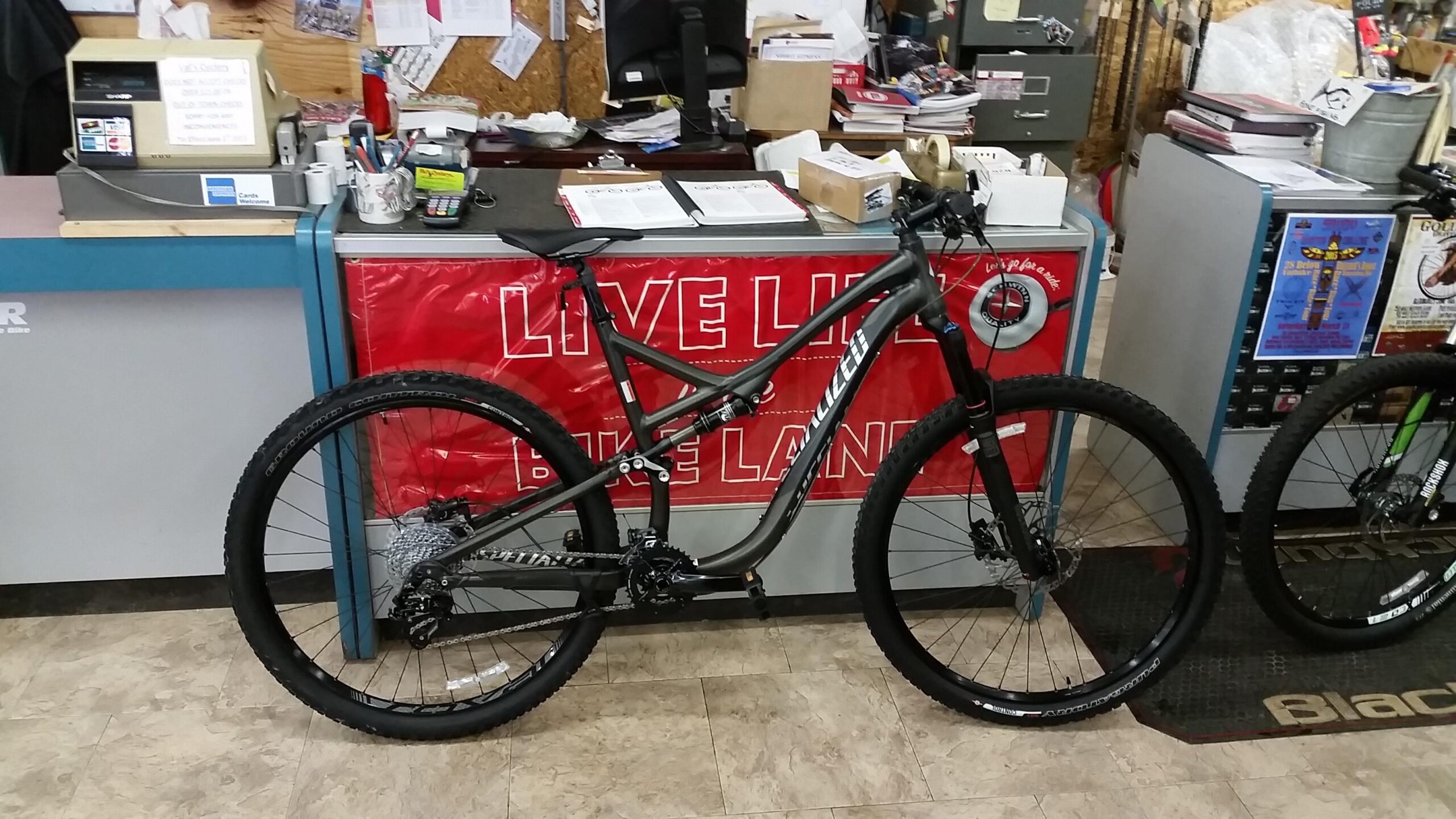 Specialized Stumpjumper FSR Comp 29er: A mountain bike is displayed in a bike shop in front of a counter covered with papers and office supplies. The bike features a sleek design with a black and gray color scheme, large wheels, and knobby tires, suitable for off-road riding. In the background, a red banner reads "LIVE LIFE BIKE LAND."