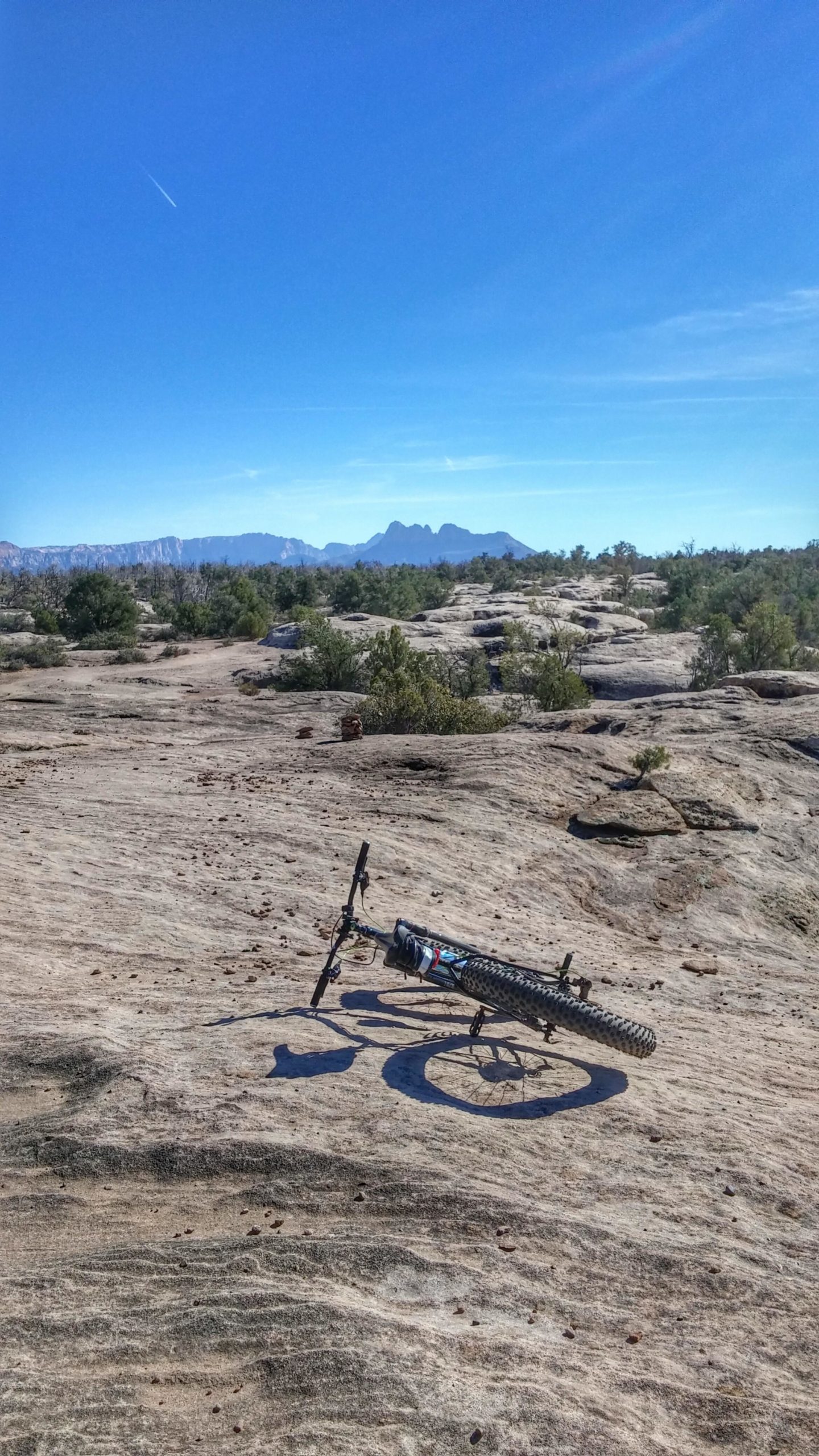 A mountain bike resting upright on a smooth, rocky surface with a clear blue sky in the background. Majestic mountains are visible in the distance, surrounded by sparse vegetation. Guacamole Mesa mountain bike trail.