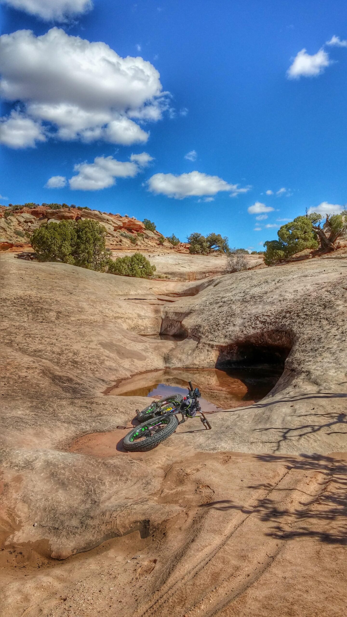 A fat bike rests on a rocky terrain with grooves and a small puddle, surrounded by desert vegetation and red rock formations under a bright blue sky with fluffy white clouds. Klondike Bluffs mountain bike trail.