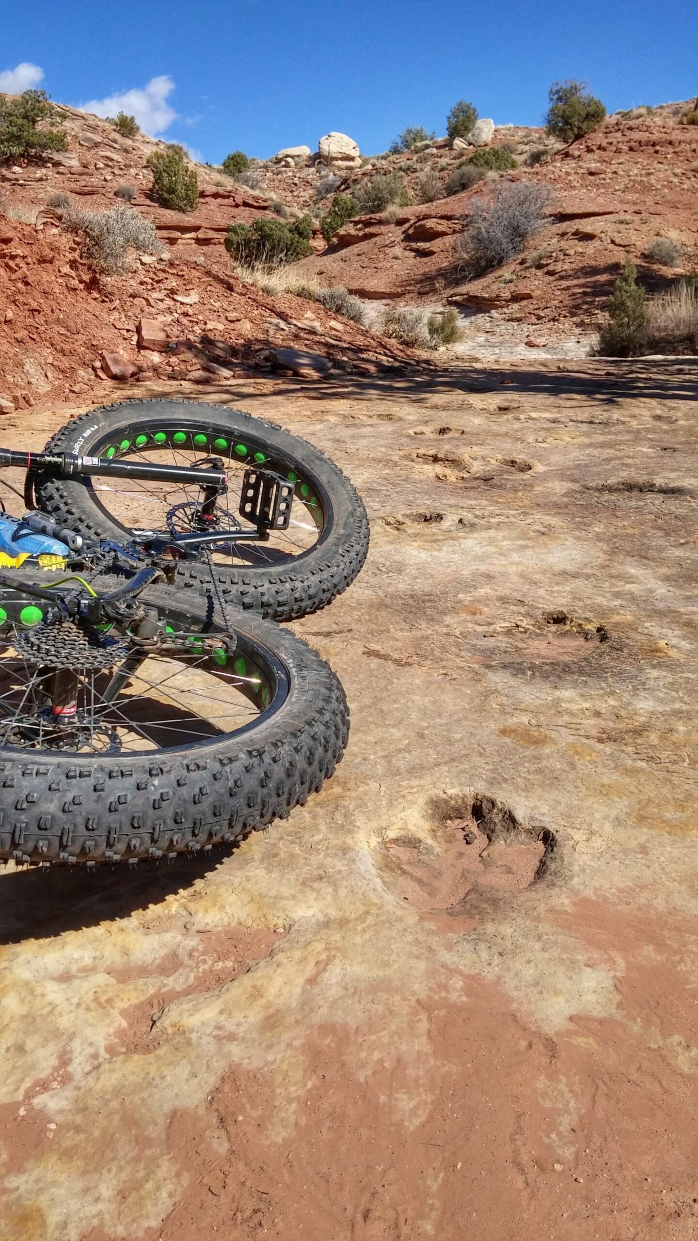 A close-up view of a fat bike with distinctive green accents resting on rocky terrain, showing large tire tracks and a nearby imprint in the ground. The background features a red rock landscape under a clear blue sky, with sparse vegetation and rugged terrain visible. Klondike Bluffs mountain bike trail.