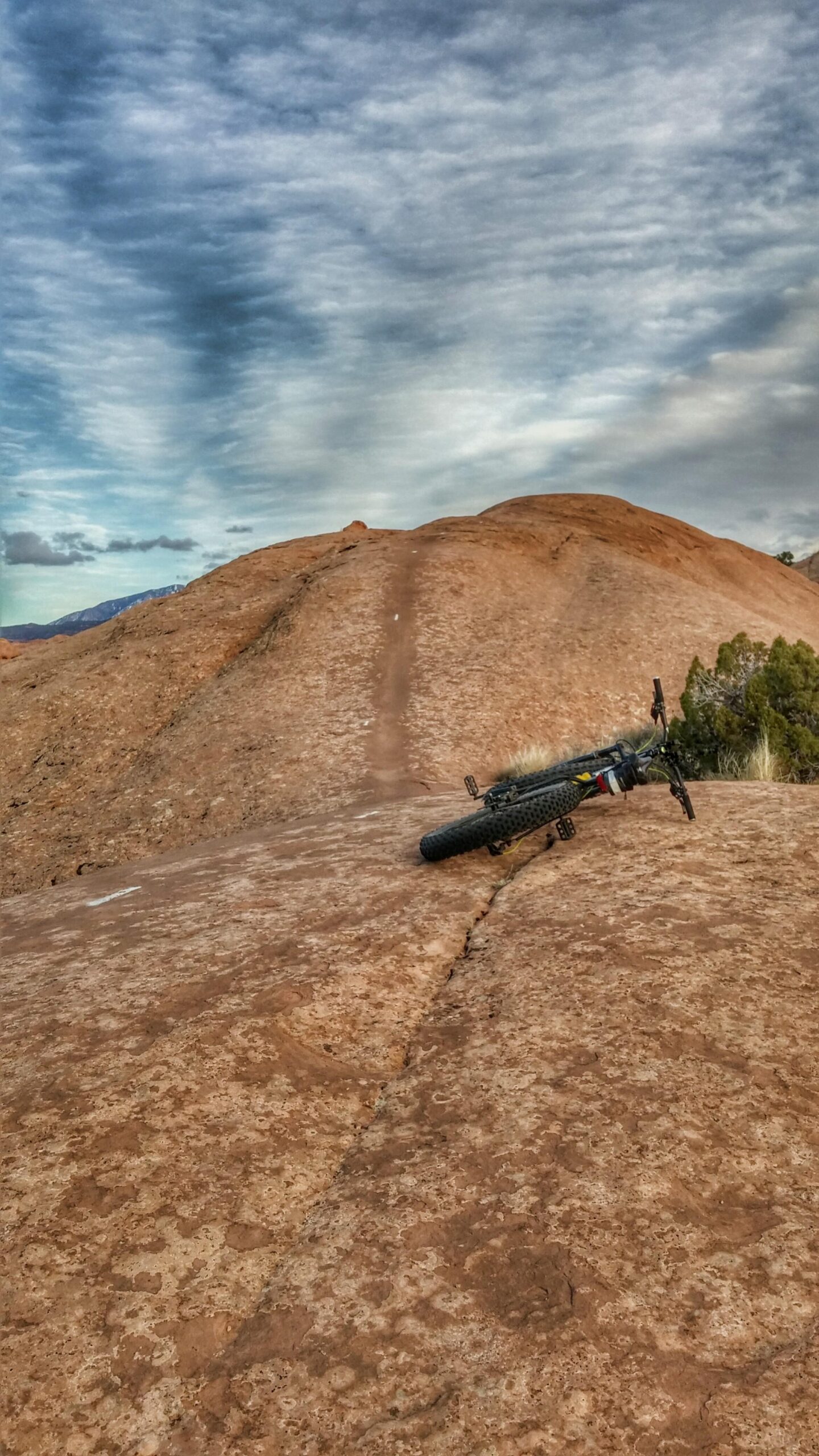 A mountain bike rests on a rocky terrain with a winding dirt path leading up a hill. The sky is filled with textured clouds, hinting at an outdoor adventure in a rugged landscape. Slickrock mountain bike trail.