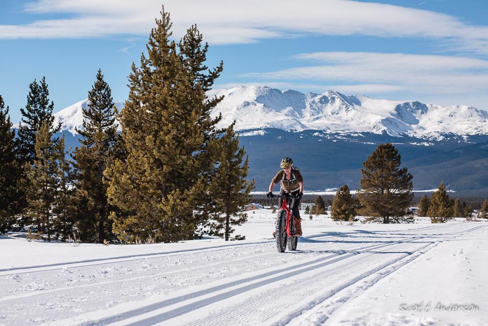 A person riding a fat bike on a snow-covered trail, surrounded by tall pine trees with snow-capped mountains in the background under a clear blue sky. Mineral Belt Trail mountain bike trail.