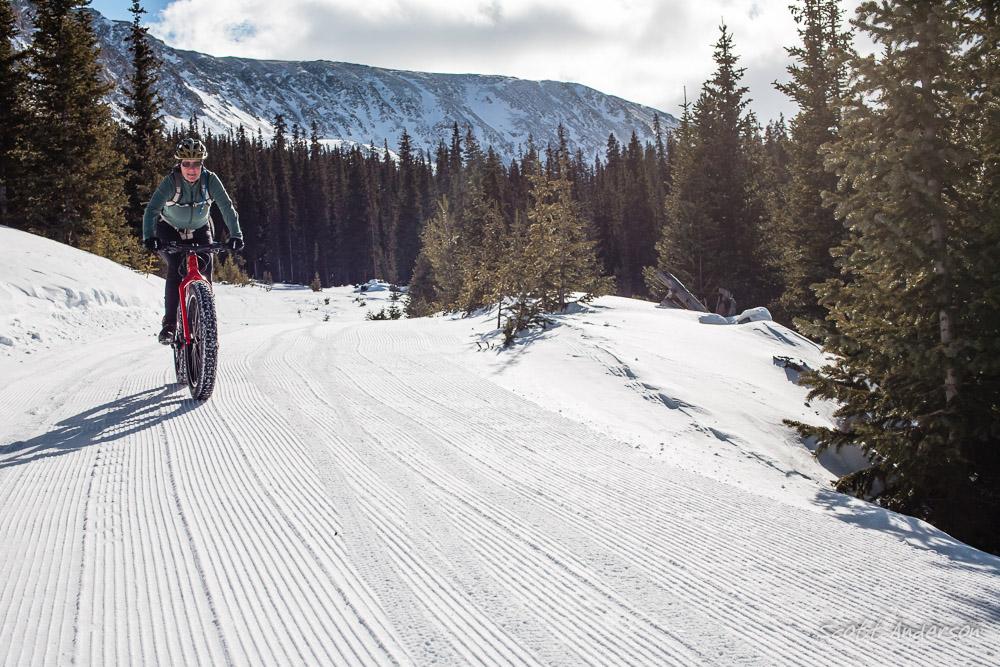 A person riding a fat tire bike on a snowy trail surrounded by evergreen trees and mountains in the background. The path shows fresh, groomed tracks in the snow, with a clear blue sky overhead. Cottonwood Pass Road mountain bike trail.