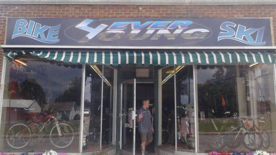 Image of the storefront of a shop named "Bike 4 Ever Young Ski." The sign features bold, colorful lettering and the entrance is framed by a green and white striped awning. Inside, bicycles are visible through the glass windows, and a person is standing near the entrance.