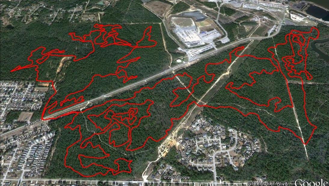 An aerial map view showing a densely wooded area with outlined trails highlighted in red. The image also includes nearby residential streets and a section of a highway. Industrial buildings are visible in the upper right corner. The layout illustrates the relationship between the natural landscape and urban development. UWF Mountain Bike Trails mountain bike trail.