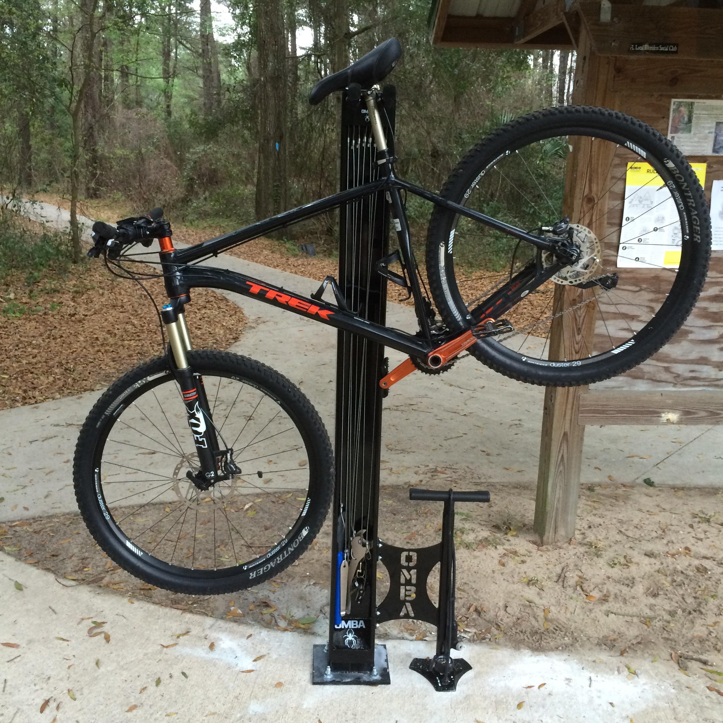 A black mountain bike with orange accents is mounted on a bike repair stand in a wooded area. The bike's front wheel is elevated, and a black floor pump is positioned next to the stand. In the background, a dirt path and wooden sign can be seen, indicating a biking trail. Santos mountain bike trail.
