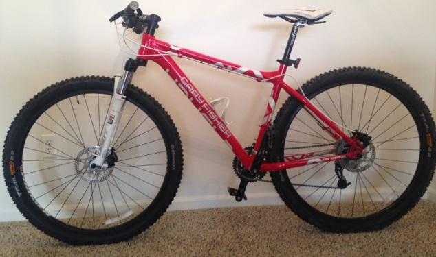 Gary Fisher Mamba: A red mountain bike with black tires, featuring a white seat and front suspension, positioned against a light-colored wall on a carpeted surface.