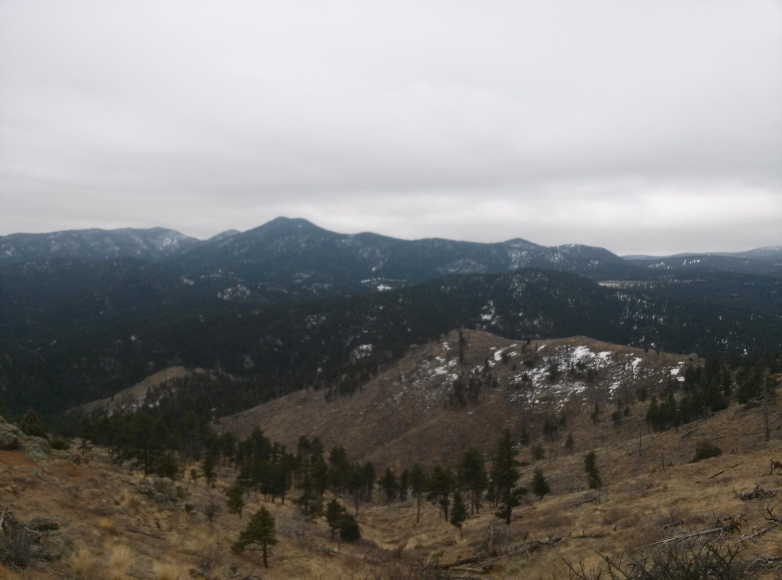 A panoramic view of a mountain landscape under a cloudy sky, featuring a range of mountains in the background and rolling hills with sparse vegetation in the foreground. Some areas are covered in snow, while evergreen trees dot the lower elevations. Walker Ranch mountain bike trail.