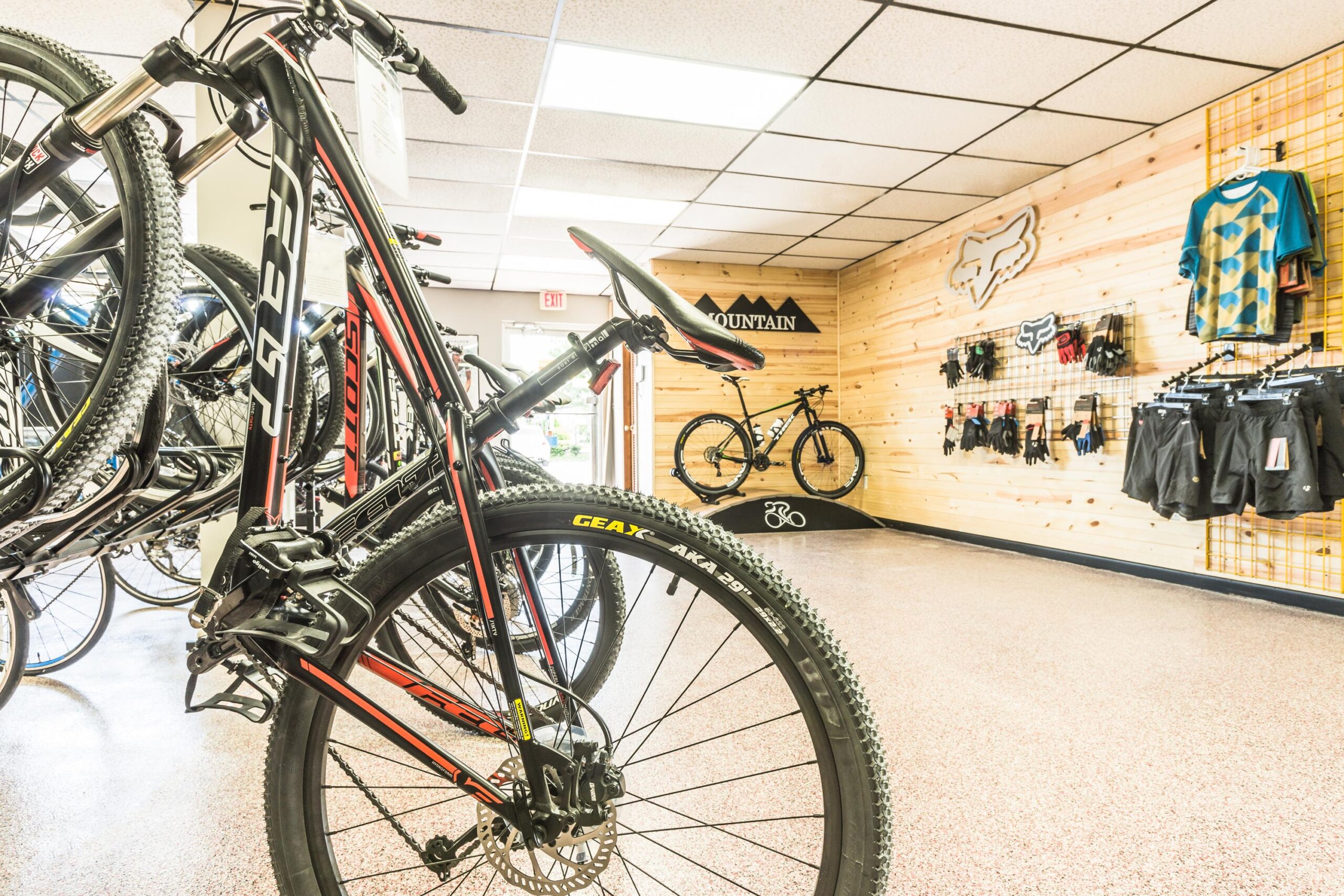 A well-lit bicycle store showcasing various mountain bikes. In the foreground, a black and red bike is displayed prominently, with additional bikes hanging on a rack in the background. The store features wooden paneling and a selection of cycling apparel and accessories on the walls, including gloves and shorts.