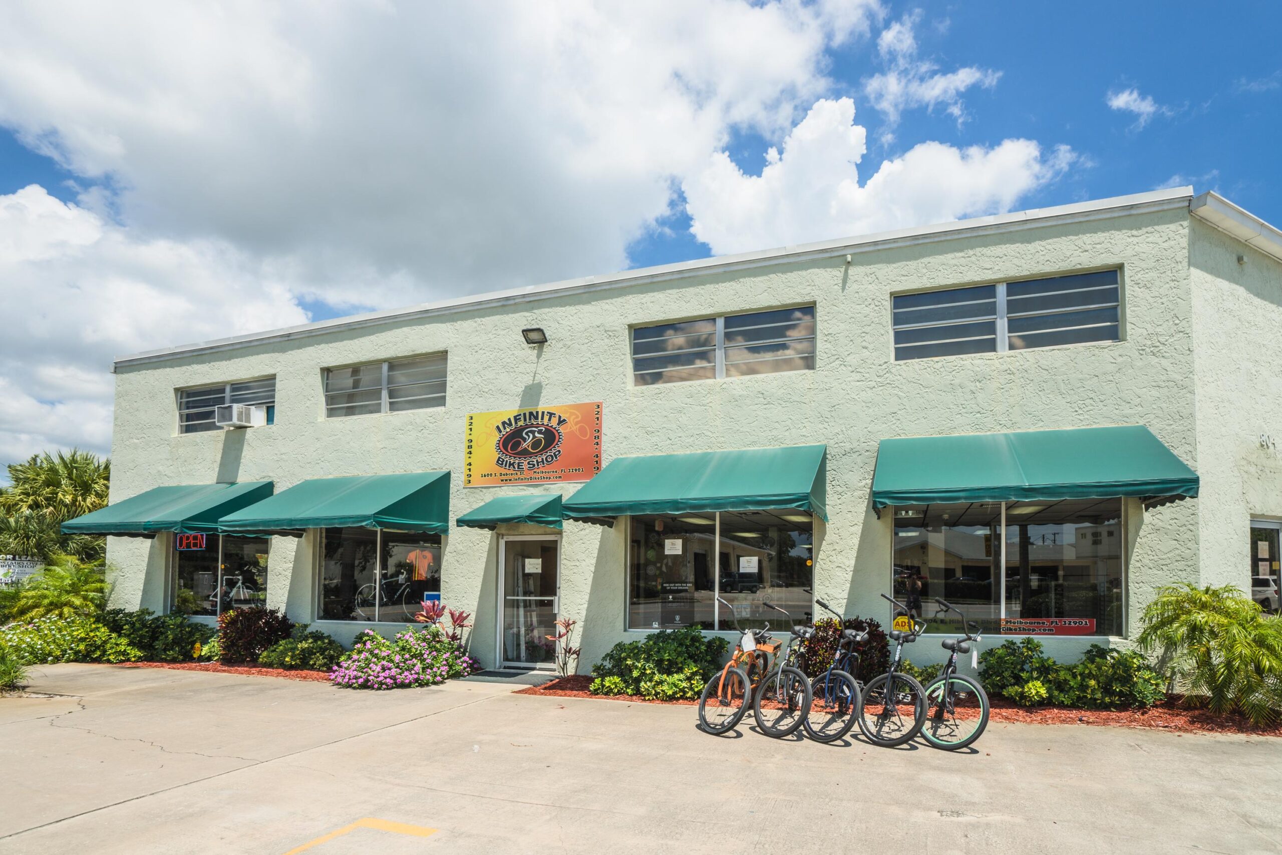 Exterior view of a bike shop named "Infinity Bike Shop," featuring a light green building with large windows, green awnings, and a bright sign. The entrance is visible, and there are several bicycles parked outside. Surrounding the shop are flowers and greenery under a partly cloudy blue sky. The "OPEN" sign is illuminated, indicating the store is welcoming customers.