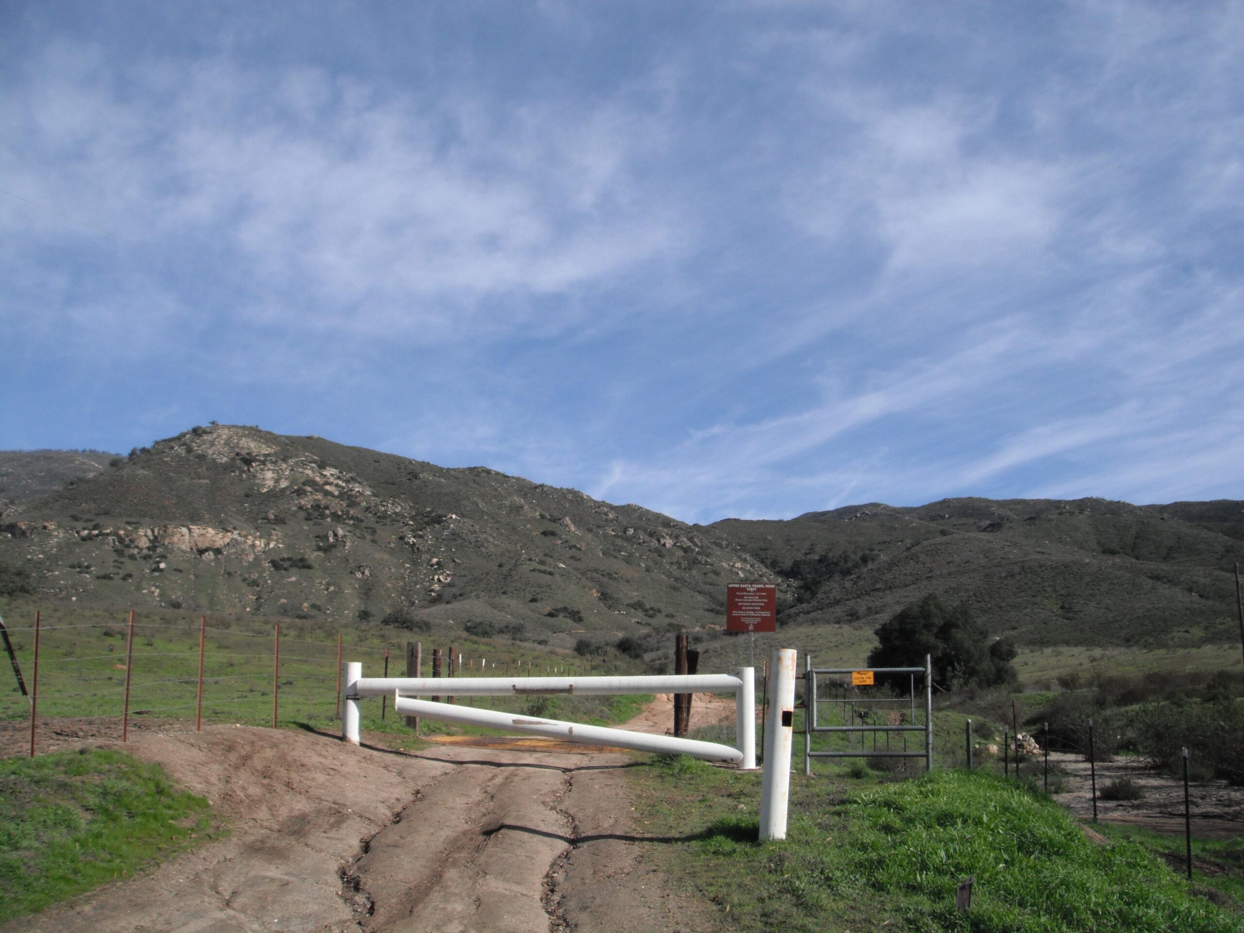 A dirt road leading to a white gate, surrounded by a wire fence and rolling green hills under a blue sky with scattered clouds. A sign is visible beside the gate, indicating regulations or warnings for the area. Upper Santa Ysabel Truck Trail mountain bike trail.