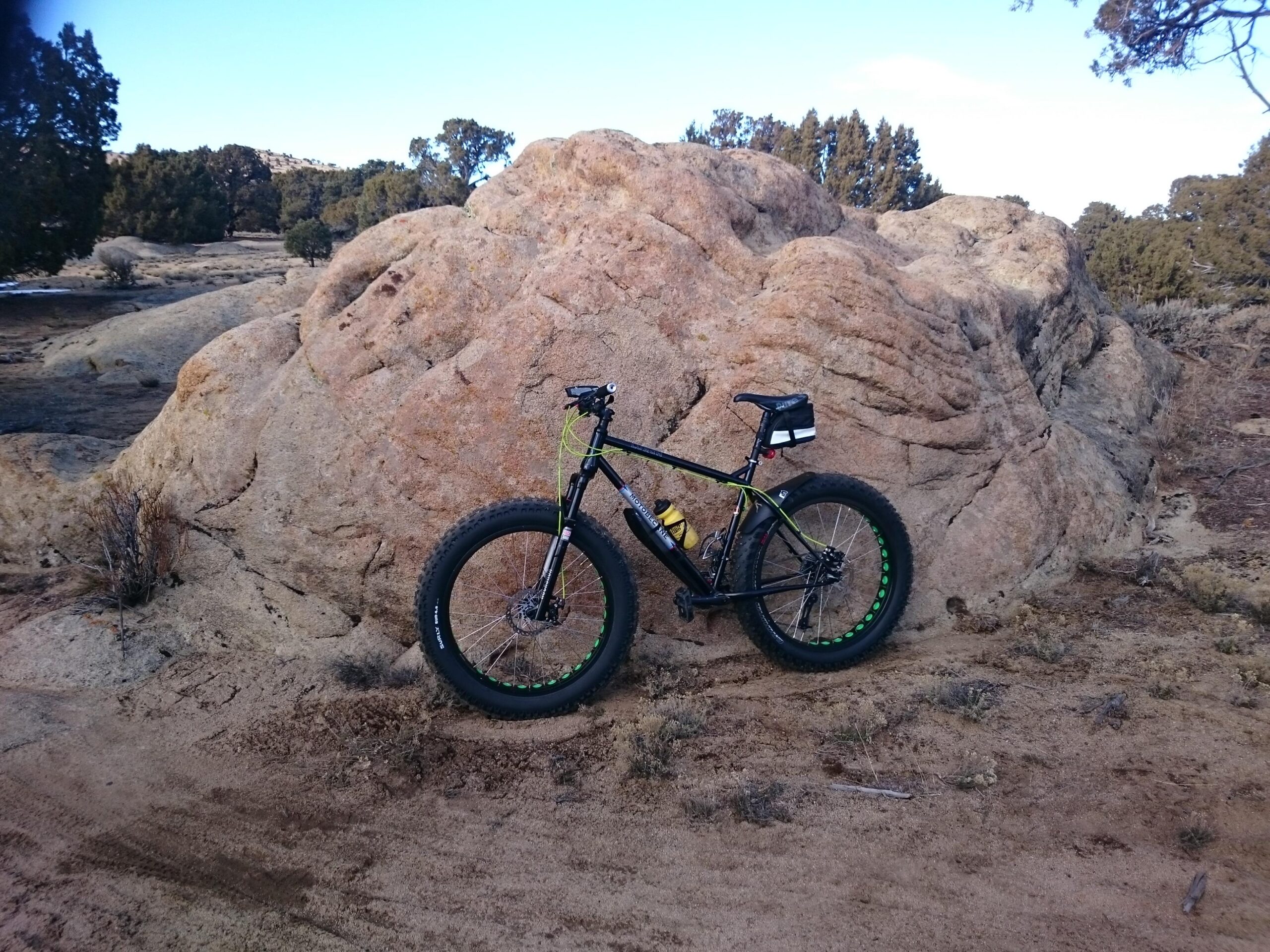 Motobecane Lurch: A fat bike parked beside a large, textured rock formation in an outdoor setting, with scattered shrubs and trees in the background under a clear blue sky.