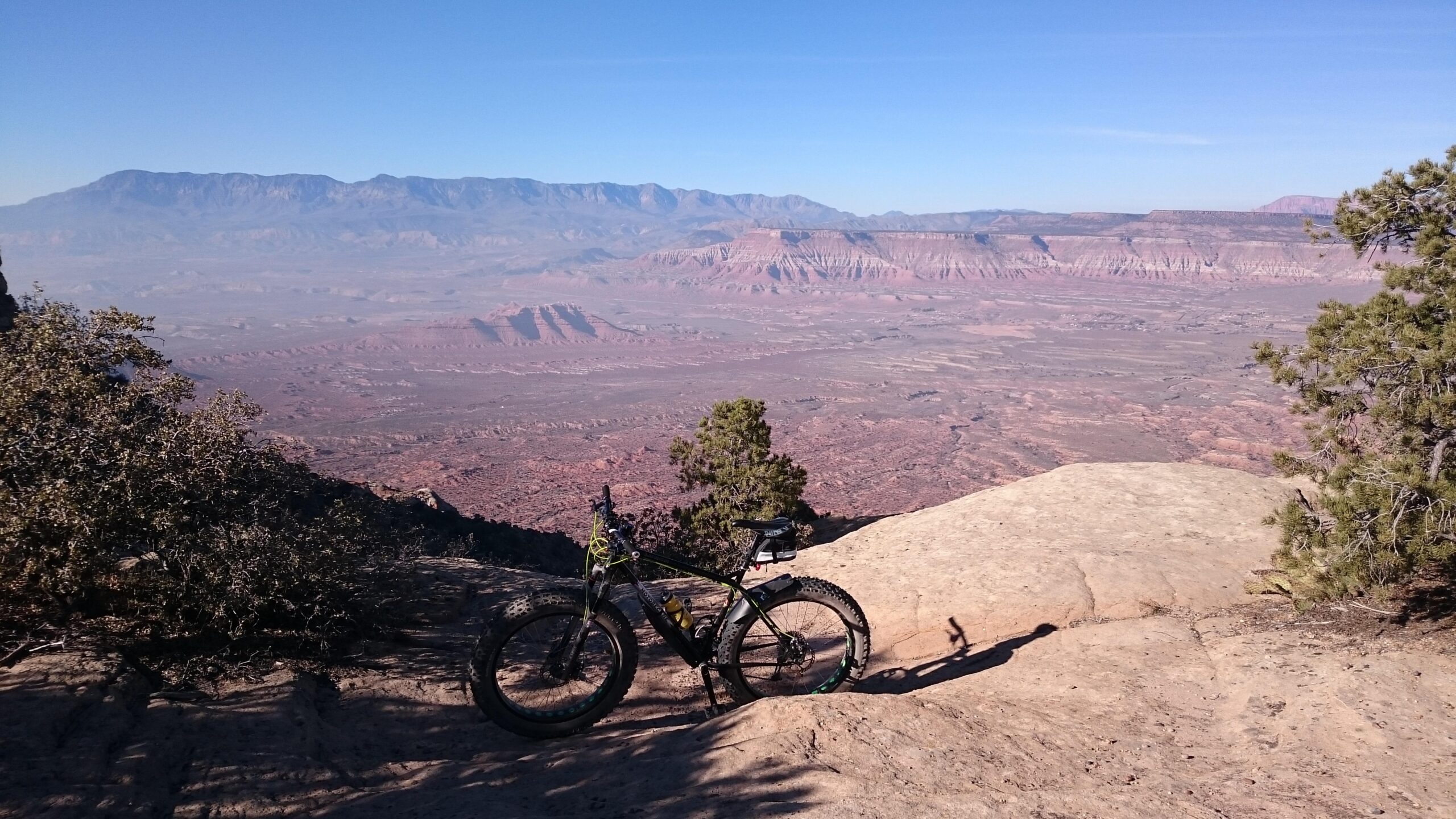 Motobecane Lurch: A mountain bike resting on a rocky outcrop with a panoramic view of layered red rock formations and distant mountains under a clear blue sky.
