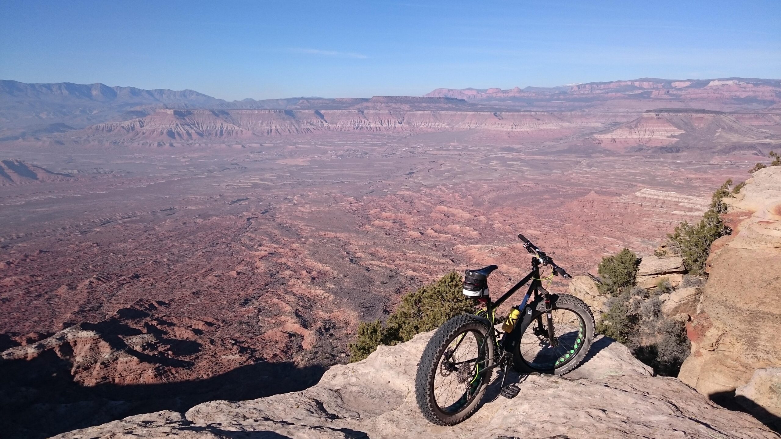 Motobecane Lurch: A black mountain bike with green accents rests on a rocky outcrop, overlooking a vast canyon landscape with layers of red and brown earth. In the background, distant mountains can be seen against a clear blue sky.