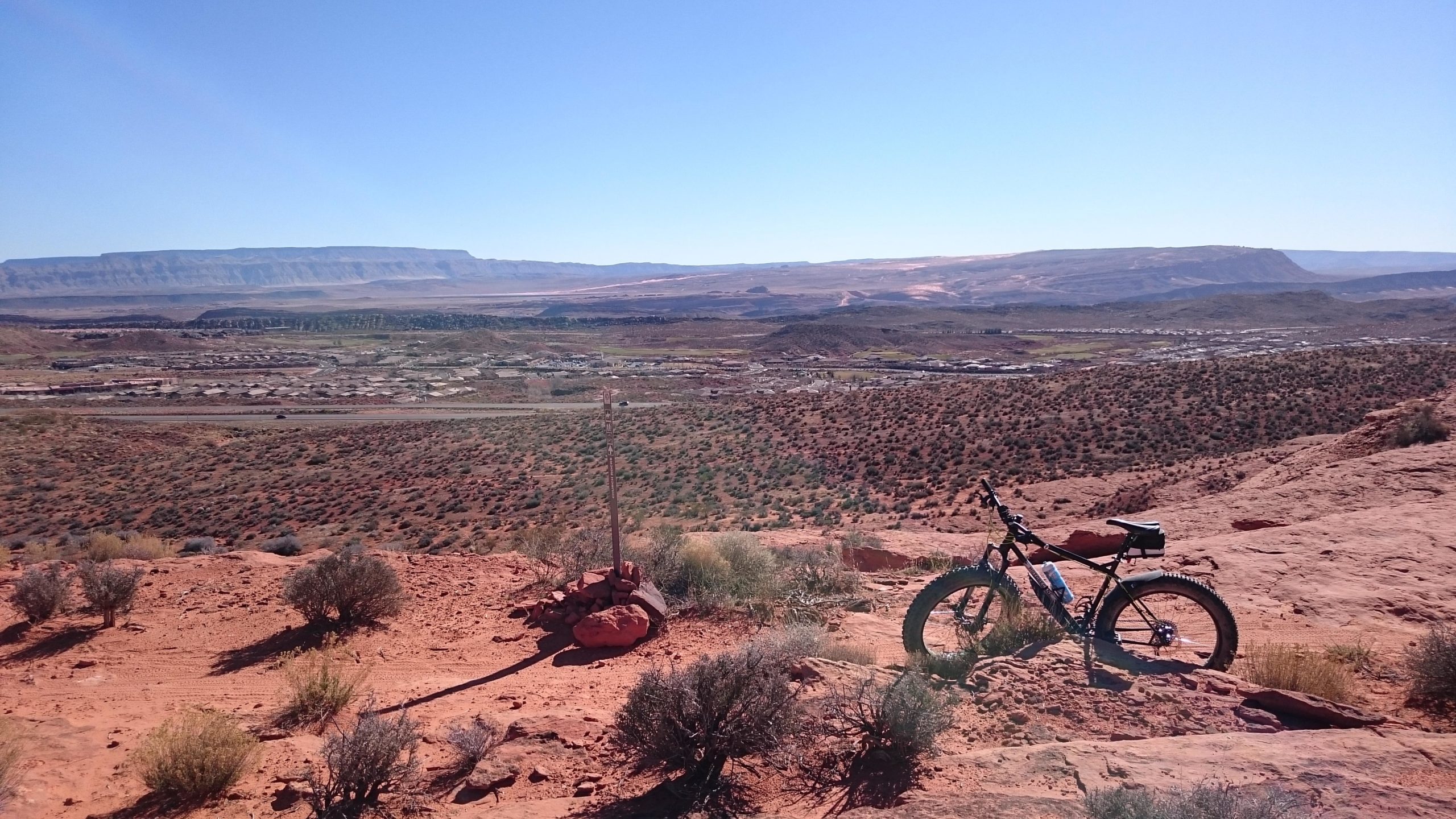 A mountain bike resting on rocky terrain, overlooking a vast desert landscape with shrubs and a distant mountain range under a clear blue sky. Prospector - Church Rocks Loop mountain bike trail.