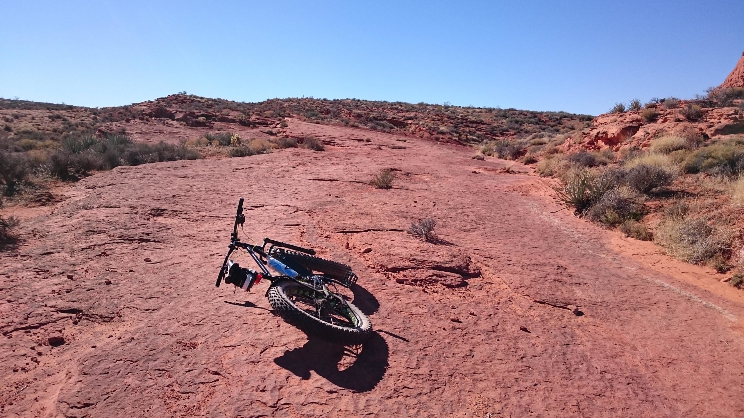 A mountain bike resting on a rocky, red terrain under a clear blue sky, with sparse vegetation in the background. Prospector - Church Rocks Loop mountain bike trail.