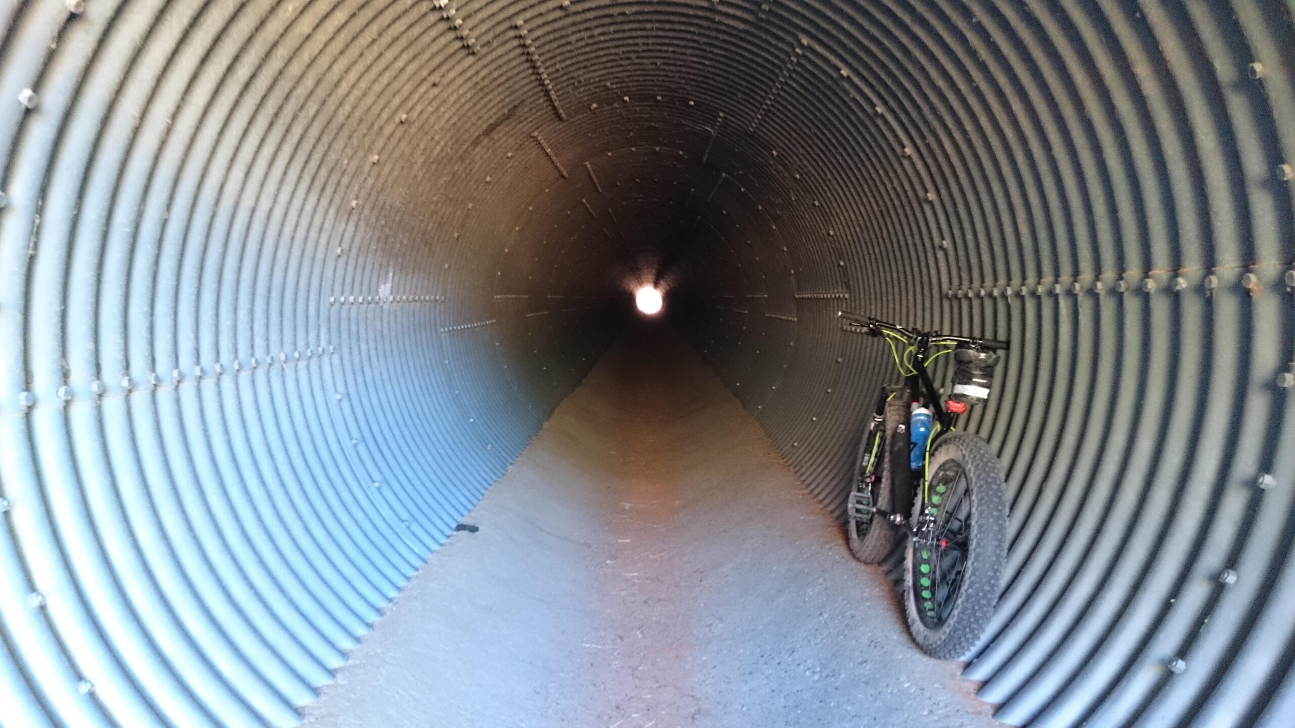 Motobecane Lurch: A bicycle is leaning against the wall inside a large, cylindrical tunnel. The tunnel features a series of horizontal ridges and is illuminated by a bright light at the far end, creating a sense of depth and perspective. The ground inside the tunnel is covered with dirt.
