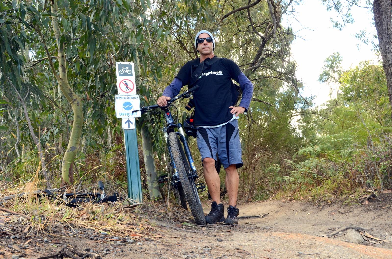 A man in a black t-shirt, shorts, and hiking boots stands next to a mountain bike on a dirt trail surrounded by trees. He is wearing sunglasses and has a backpack. Nearby, there are trail signs indicating a bike path and a difficult trail named "Upper Blair Witch." Lysterfield Mountain Bike Area mountain bike trail.