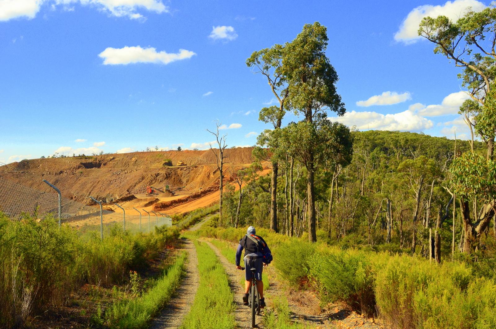 A cyclist rides along a gravel path bordered by greenery, with a hilly landscape and blue sky in the background. In the distance, a construction site is visible, featuring machinery and excavated earth, surrounded by a fence. Lysterfield Mountain Bike Area mountain bike trail.