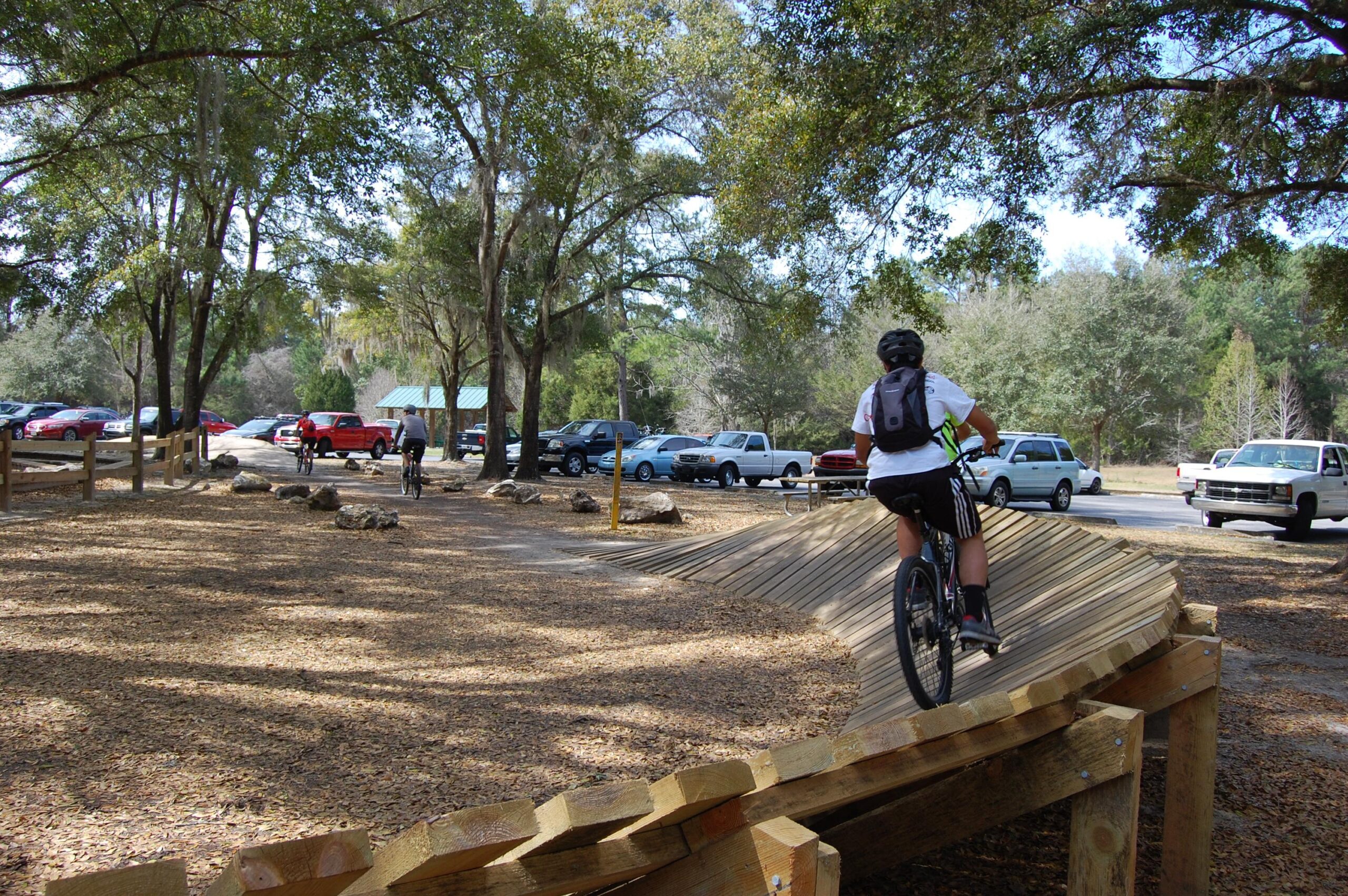 A cyclist rides along a winding wooden path in a park, surrounded by trees and parked cars in the background. The pathway is designed for biking, with a smooth surface and gentle curves, while other cyclists can be seen in the distance. The area is covered in leaves, creating a natural setting for outdoor activities. Santos mountain bike trail.