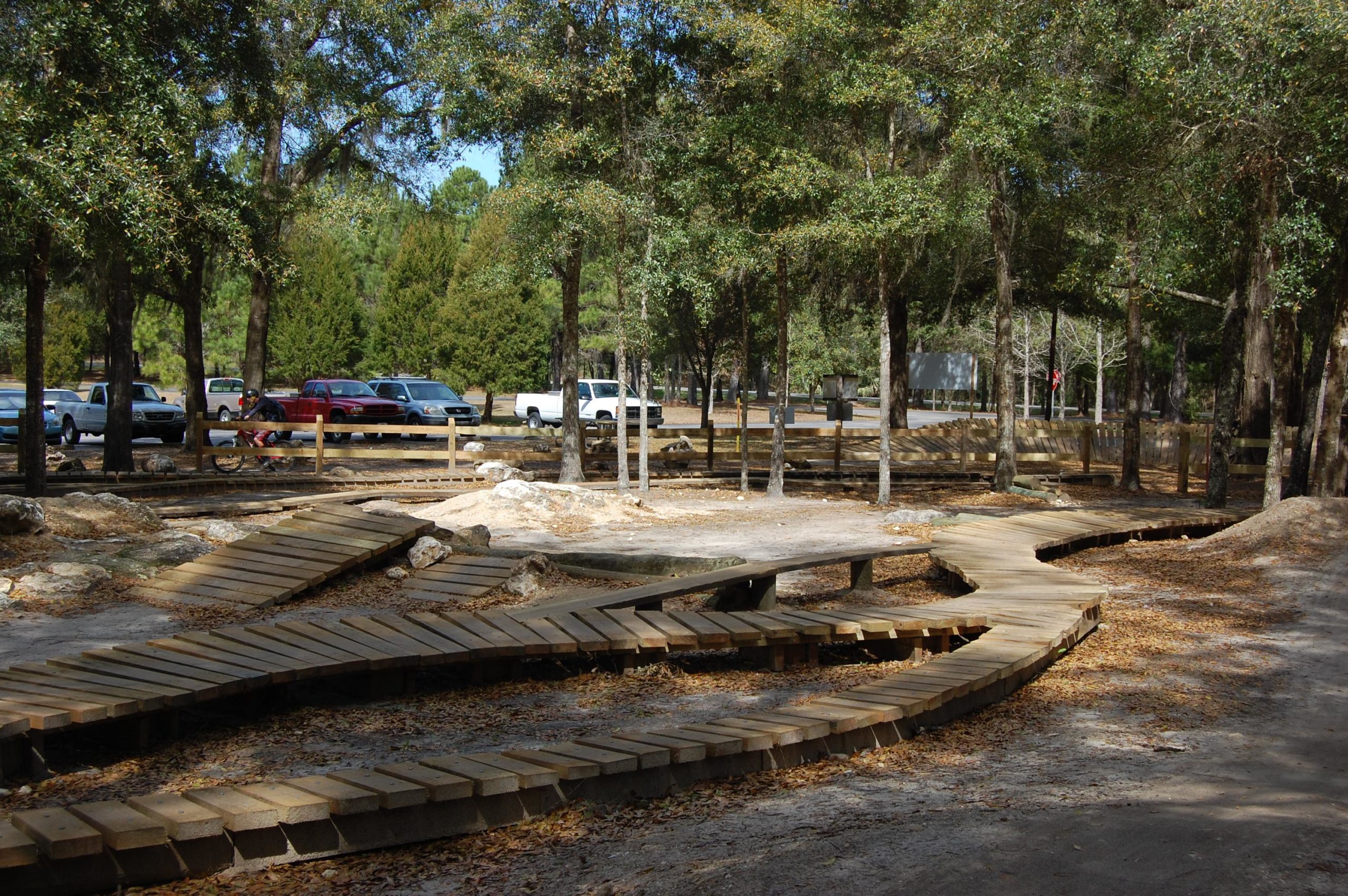 A winding wooden walkway covered with leaves, surrounded by trees, leads through a park area. In the background, several parked vehicles are visible among the trees, with additional wooden structures hinting at recreational facilities nearby. The scene is bright and inviting, showcasing a natural, wooded environment. Santos mountain bike trail.