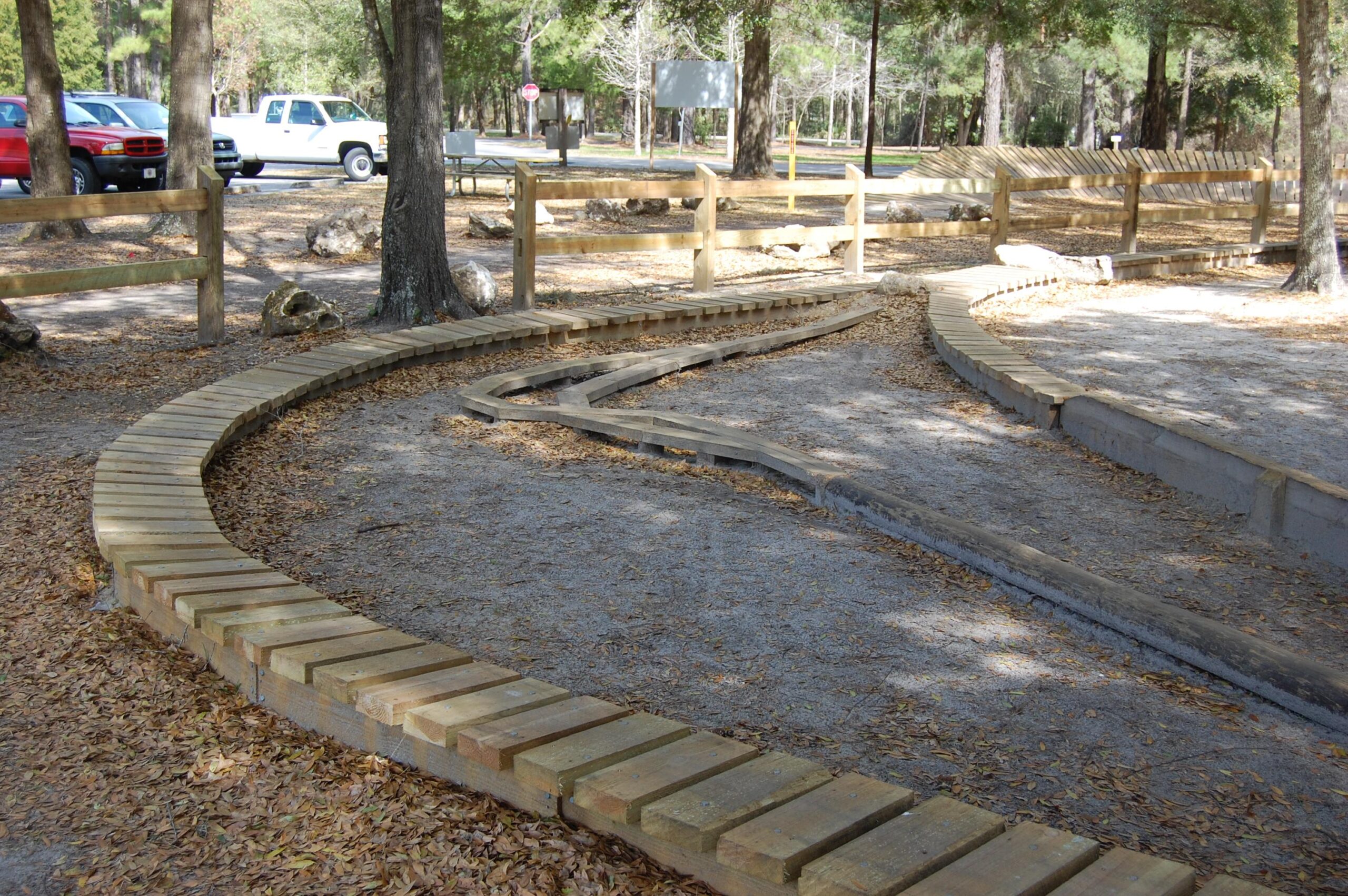 A winding wooden path surrounded by trees and leaf-covered ground, leading through a park area. The pathway includes sections made from planks of wood, creating a natural trail, with gravel in the center. In the background, parked vehicles and signs are visible along a dirt road. Santos mountain bike trail.