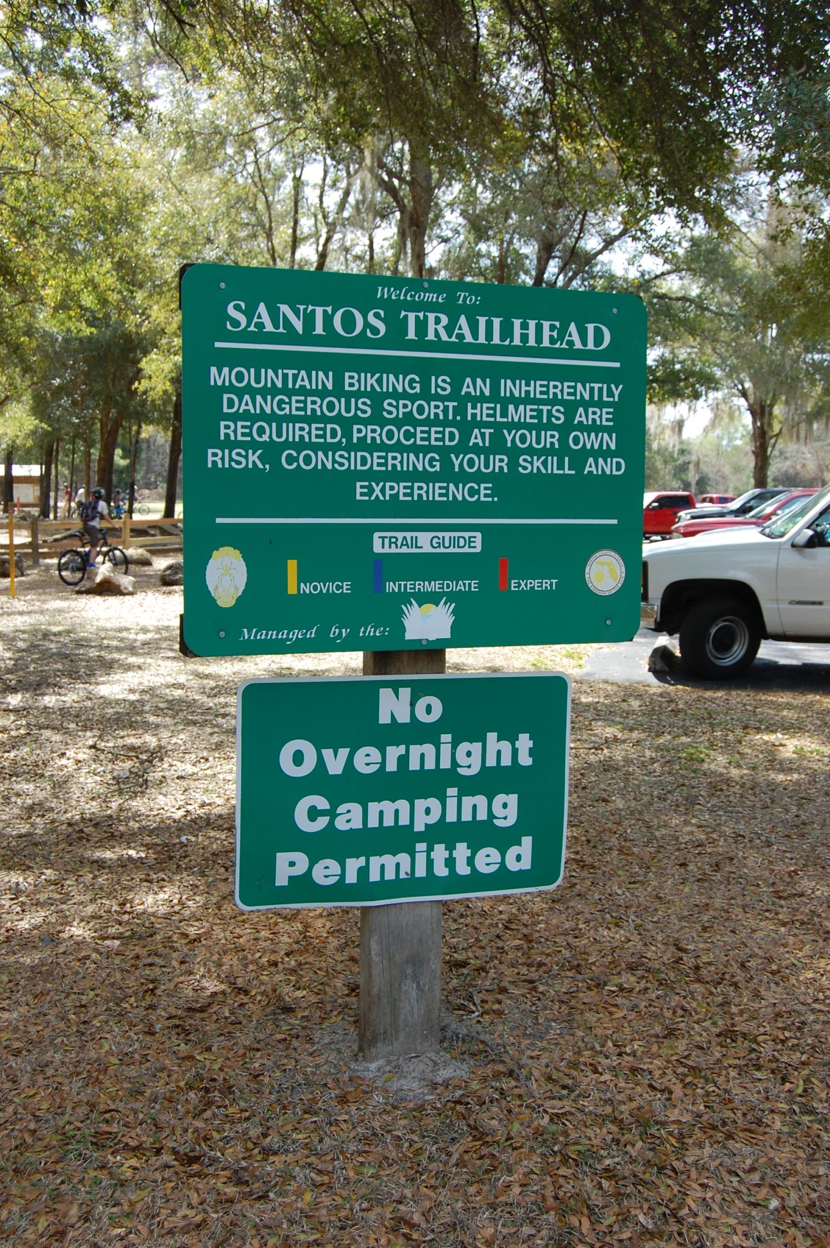 A green sign welcoming visitors to Santos Trailhead, providing safety information about mountain biking. The sign states that helmets are required and emphasizes the inherent dangers of the sport. Below, a trail guide categorizes trails as Novice, Intermediate, and Expert. A second sign notes that overnight camping is not permitted. The background features trees and a parking area. Santos mountain bike trail.