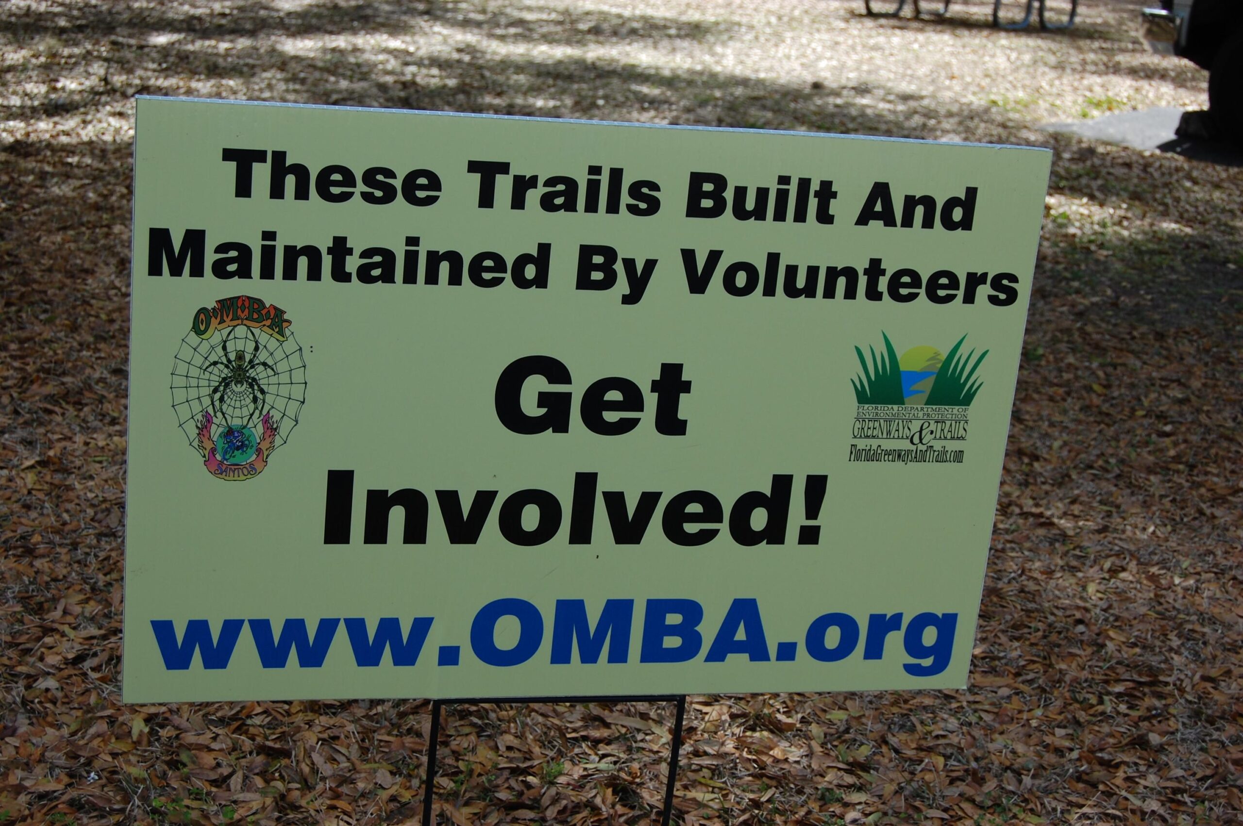 A sign promoting volunteer involvement in trail maintenance, featuring text that reads, "These Trails Built And Maintained By Volunteers. Get Involved!" It includes a logo on the left and a URL, "www.OMBA.org," at the bottom. The background consists of fallen leaves and natural scenery. Santos mountain bike trail.