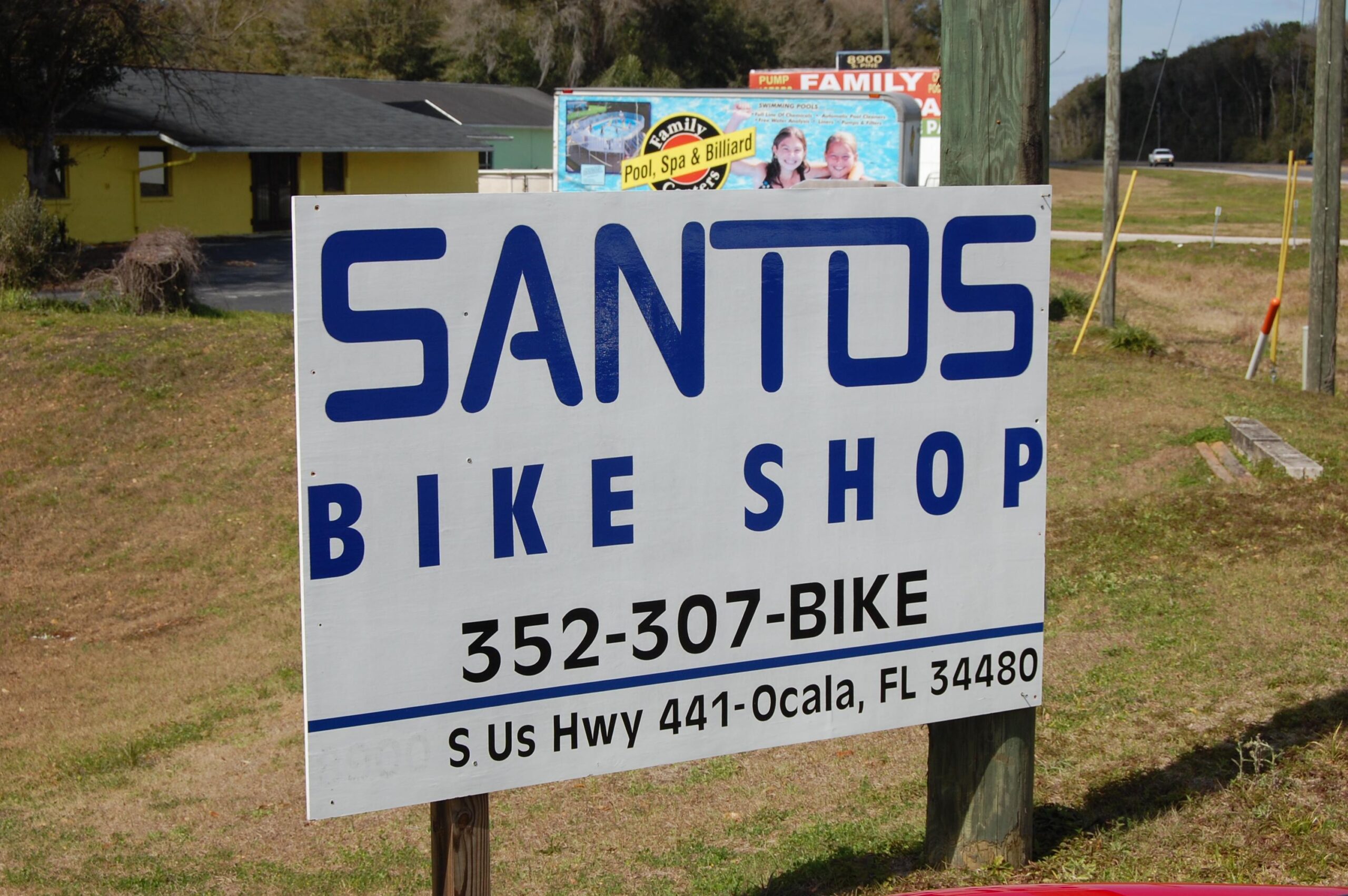 Sign for Santos Bike Shop, displaying the shop name in large blue letters, along with a contact phone number (352-307-BIKE) and the address (S. US Hwy 441, Ocala, FL 34480). The background features grassy landscaping and nearby buildings.