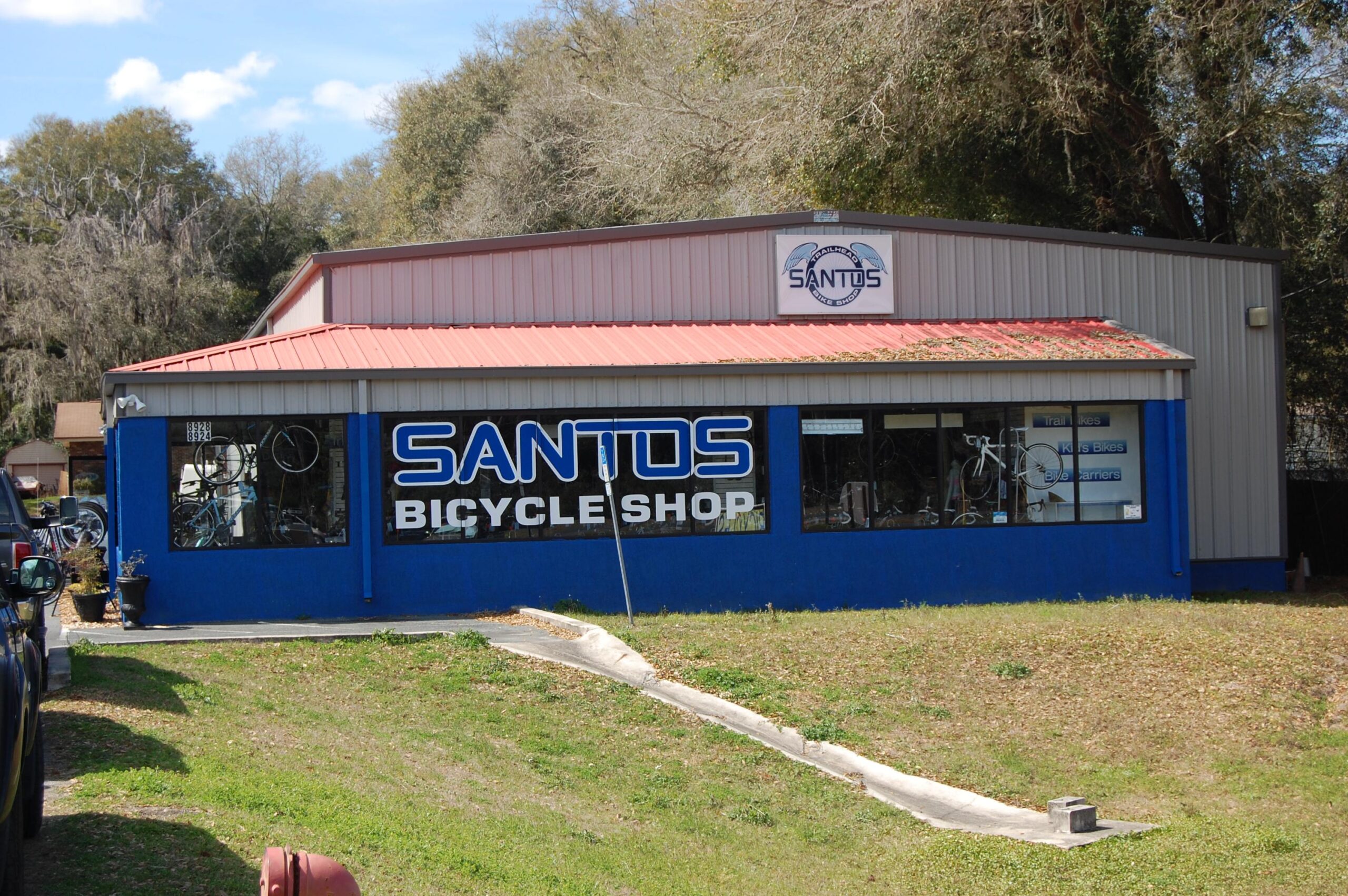 Exterior view of the Santos Bicycle Shop, featuring a blue and gray building with a red metal roof. Large windows display various bicycles and accessories. The shop's name is prominently displayed in bold white and blue letters on the front. Surrounding the building is a grassy area with some trees in the background.