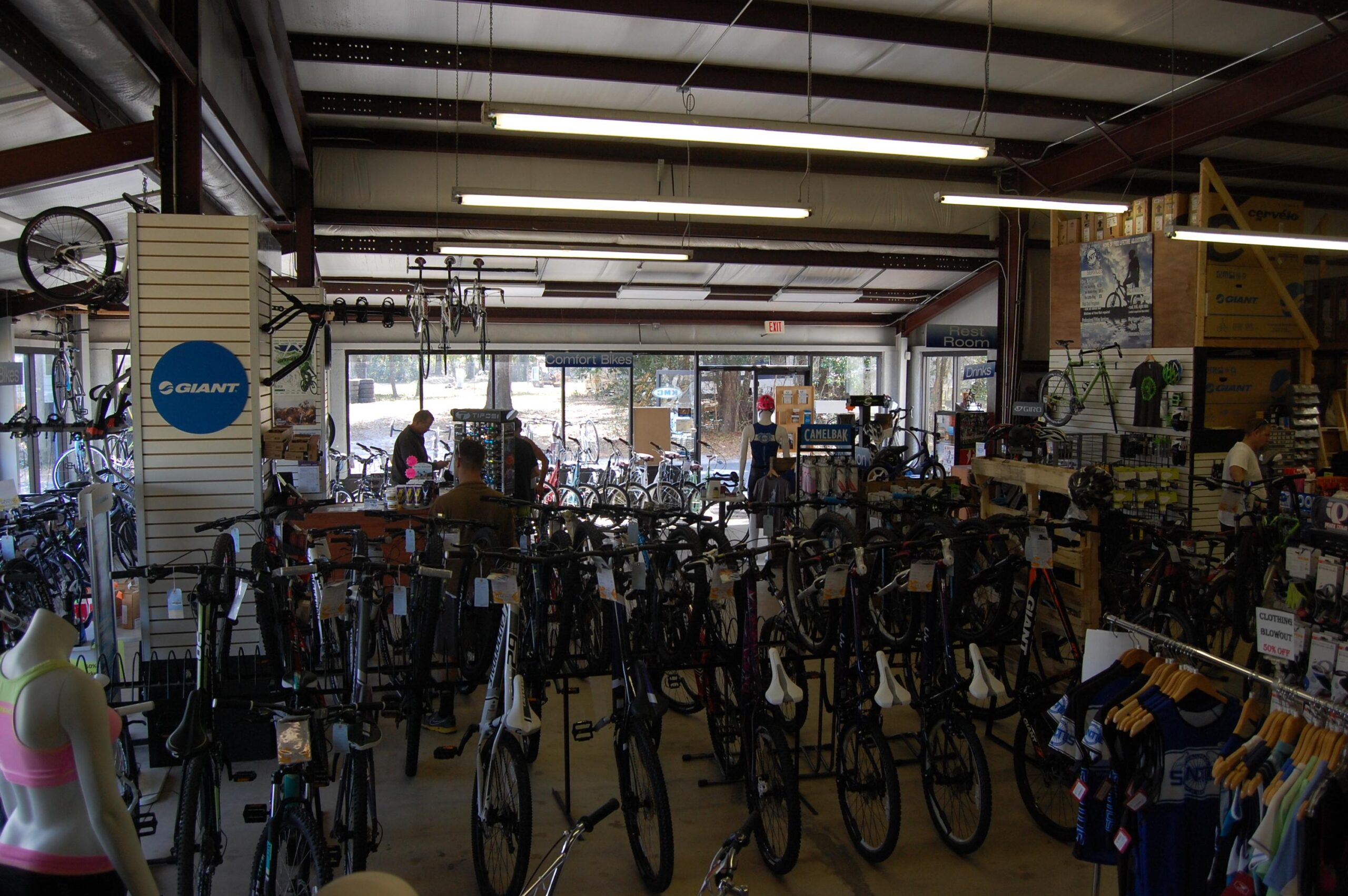 Image of a bicycle shop interior filled with various bicycles on display. The space features a central aisle with different bike models, a clothing rack on the left, and a customer service area at the back. Large windows allow natural light, and promotional signage from the brand "Giant" is visible. Customers can be seen browsing the merchandise.