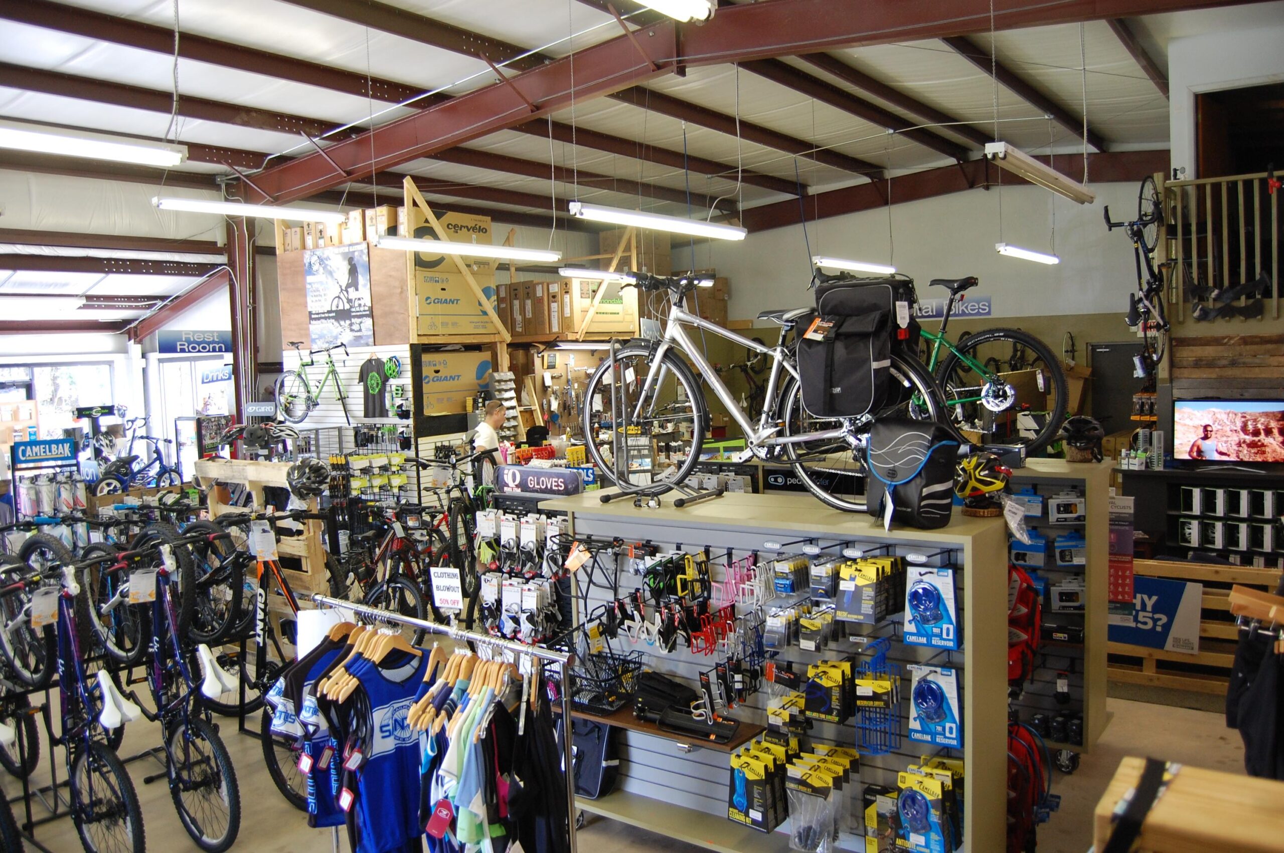Interior of a bicycle shop showcasing a variety of bikes and cycling accessories. Several bicycles are displayed prominently on shelves, with additional bikes in the background. The shop features racks filled with helmets, gloves, and other cycling gear, along with merchandise organized on wooden and metal displays. Bright lighting illuminates the space, creating an inviting shopping atmosphere.
