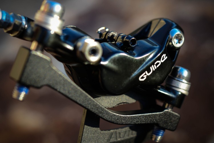 Close-up of a black bicycle brake caliper with the word "GUIDE" visible, showcasing its mechanical components and connections. The background is softly blurred, emphasizing the details of the brake part.