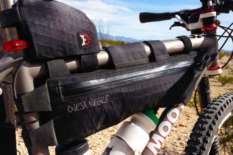 A close-up view of a mountain bike frame featuring a black bike bag with the text "Oveja Negra" on the side. The bike's handlebars, gear shifters, and tires are partially visible, set against a backdrop of blue skies and desert terrain. The bag is secured with straps to the bike frame.