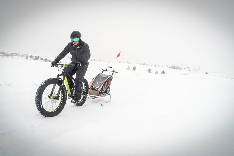 A person riding a fat bike through a snowy landscape, pulling a small sled behind them. The scene features falling snow, a thick white blanket covering the ground, and a few distant trees. The rider is dressed in winter gear, including a helmet and goggles, and the visibility is low due to the snowy conditions.