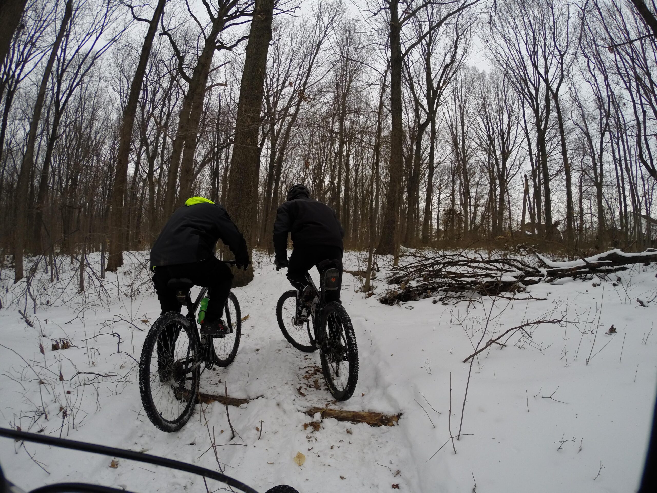 Two mountain bikers navigate a snowy trail in a wooded area, surrounded by bare trees. One biker wears a black jacket with a bright green hood, while the other is dressed entirely in black. Snow covers the ground, creating a wintery scene, as the bikers make their way along a narrow path. Trails seperated by streets mountain bike trail.