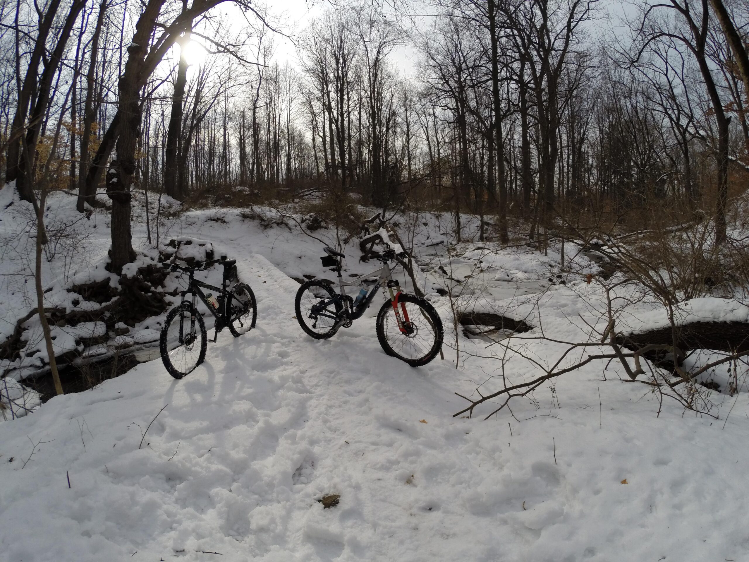 Two mountain bikes are parked on a snowy trail in a wooded area. The landscape features leafless trees and a partially cloudy sky, with sunlight filtering through the branches. The ground is covered in a thick layer of snow, and there are small streams visible in the background, adding to the winter scenery. Richmond Avenue and Forest Hill road mountain bike trail.