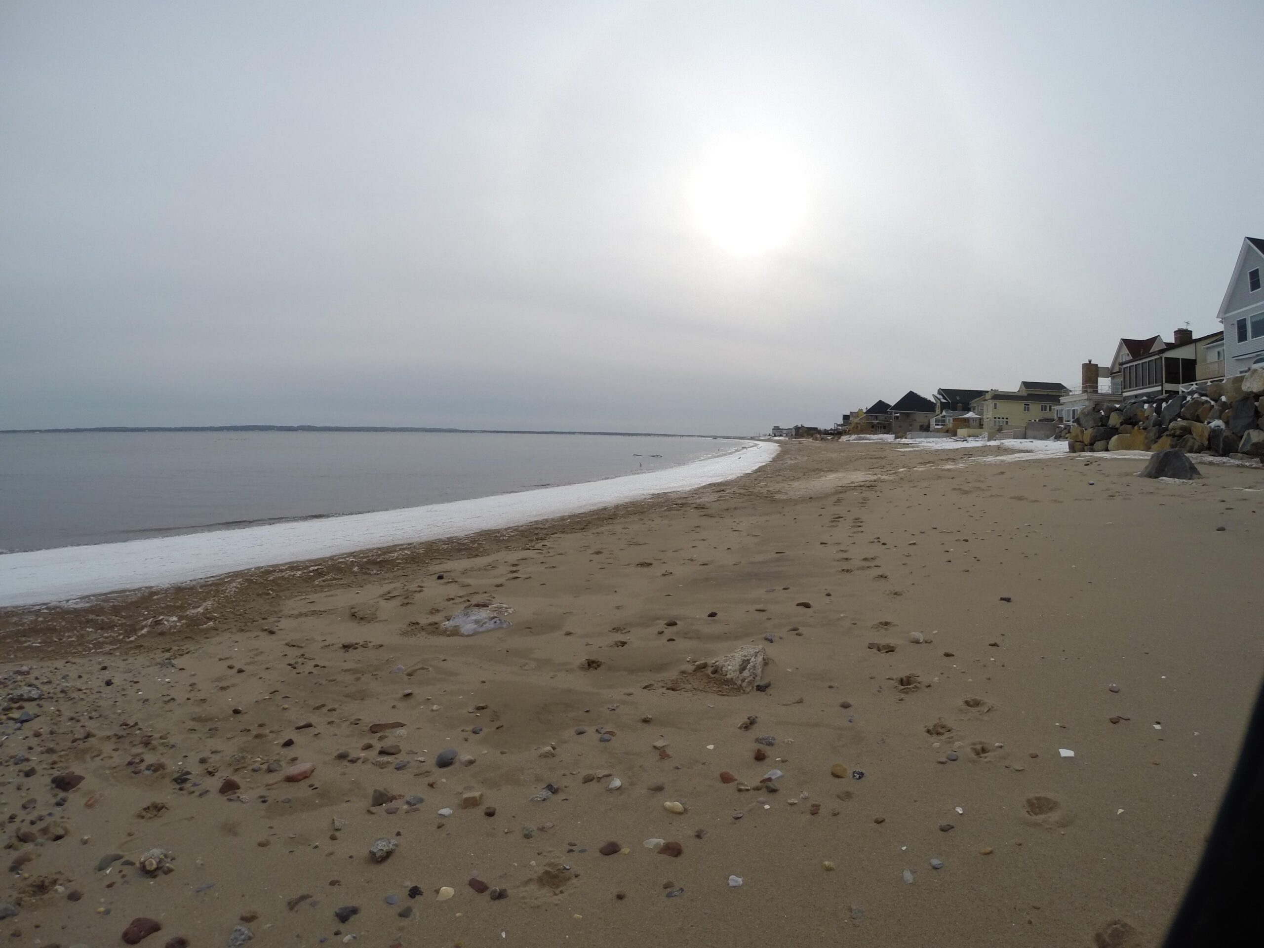 A serene beach scene featuring a sandy shoreline with scattered pebbles, a calm water surface, and a gray, overcast sky. The sun is partially visible, casting a soft light over the landscape. In the background, several houses line the beach, with rocks positioned along the water's edge. Staten Island Beach mountain bike trail.
