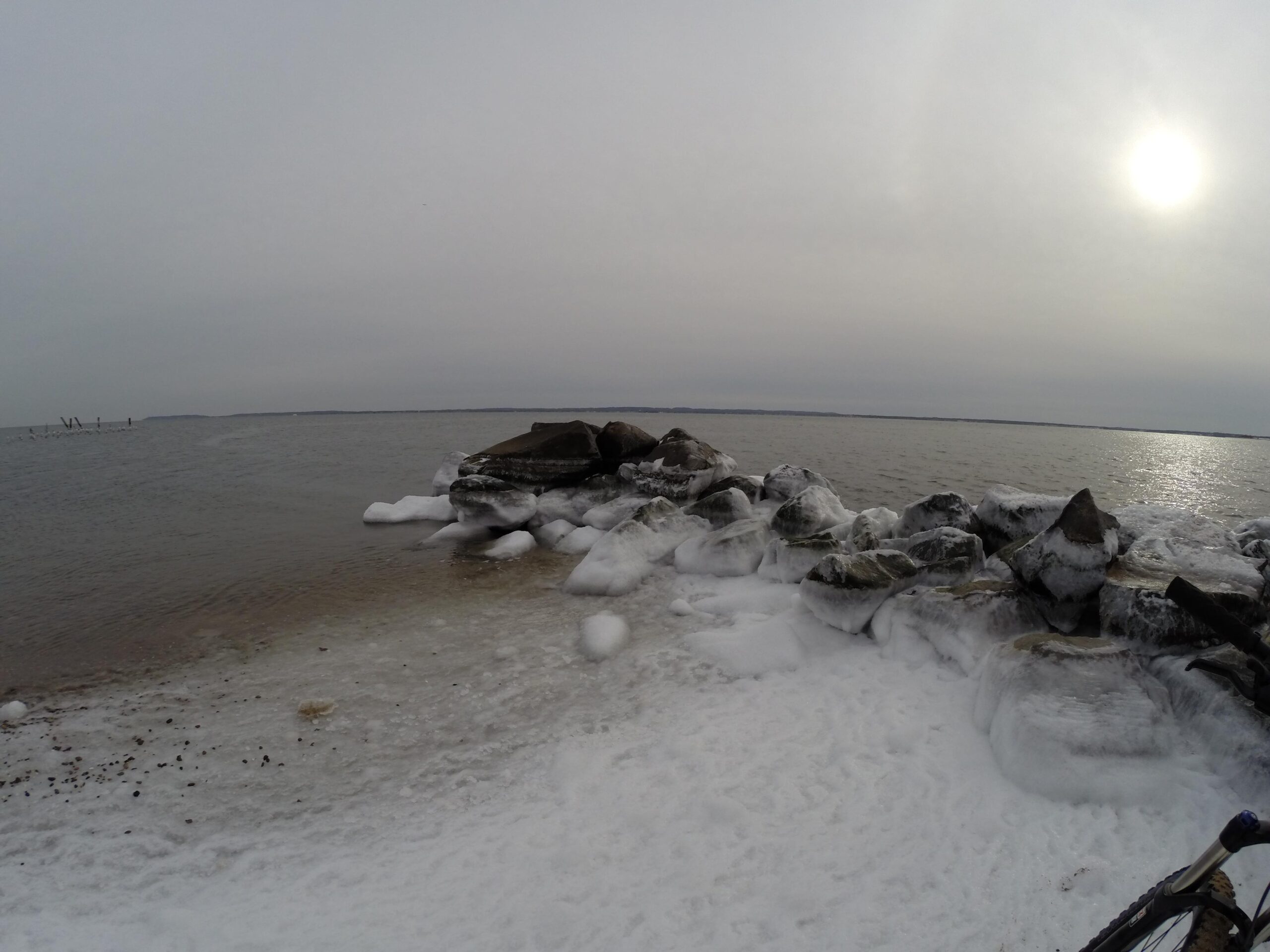 A serene winter seascape featuring icy rocks along the shoreline. The water is calm and partially frozen, with a light fog limiting visibility in the distance. The sun hangs low in a cloudy sky, casting a muted light over the scene. Snow and ice cover the rocks, creating a tranquil and chilly atmosphere. Staten Island Beach mountain bike trail.