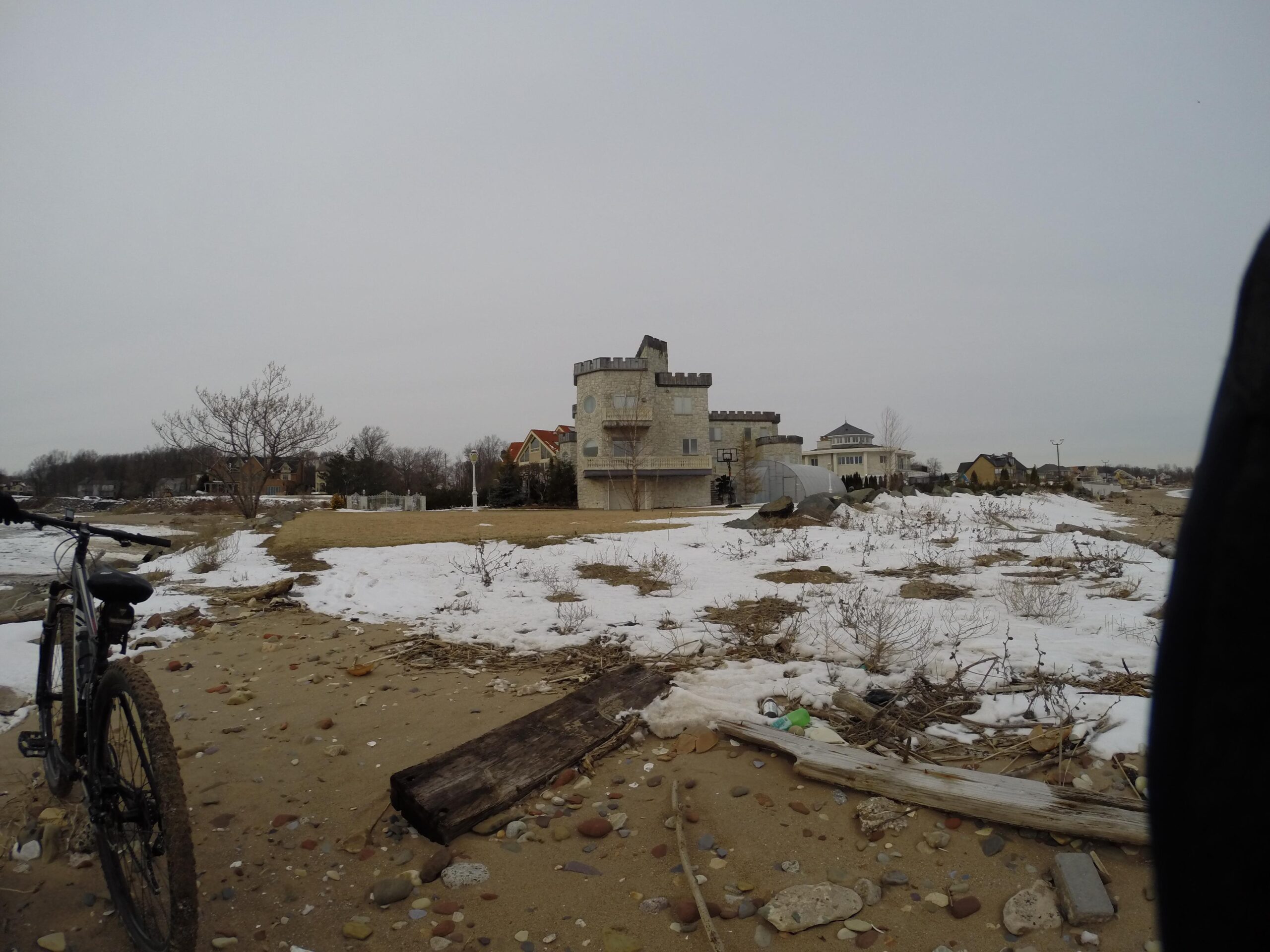 A bike is parked on a sandy beach area covered with small stones and patches of snow. In the background, there is a distinct castle-like structure made of light-colored stone, along with other houses visible behind it. The scene is set under a cloudy sky, creating a moody atmosphere. The landscape appears to be quiet and somewhat desolate, with scattered debris and sparse vegetation. Staten Island Beach mountain bike trail.